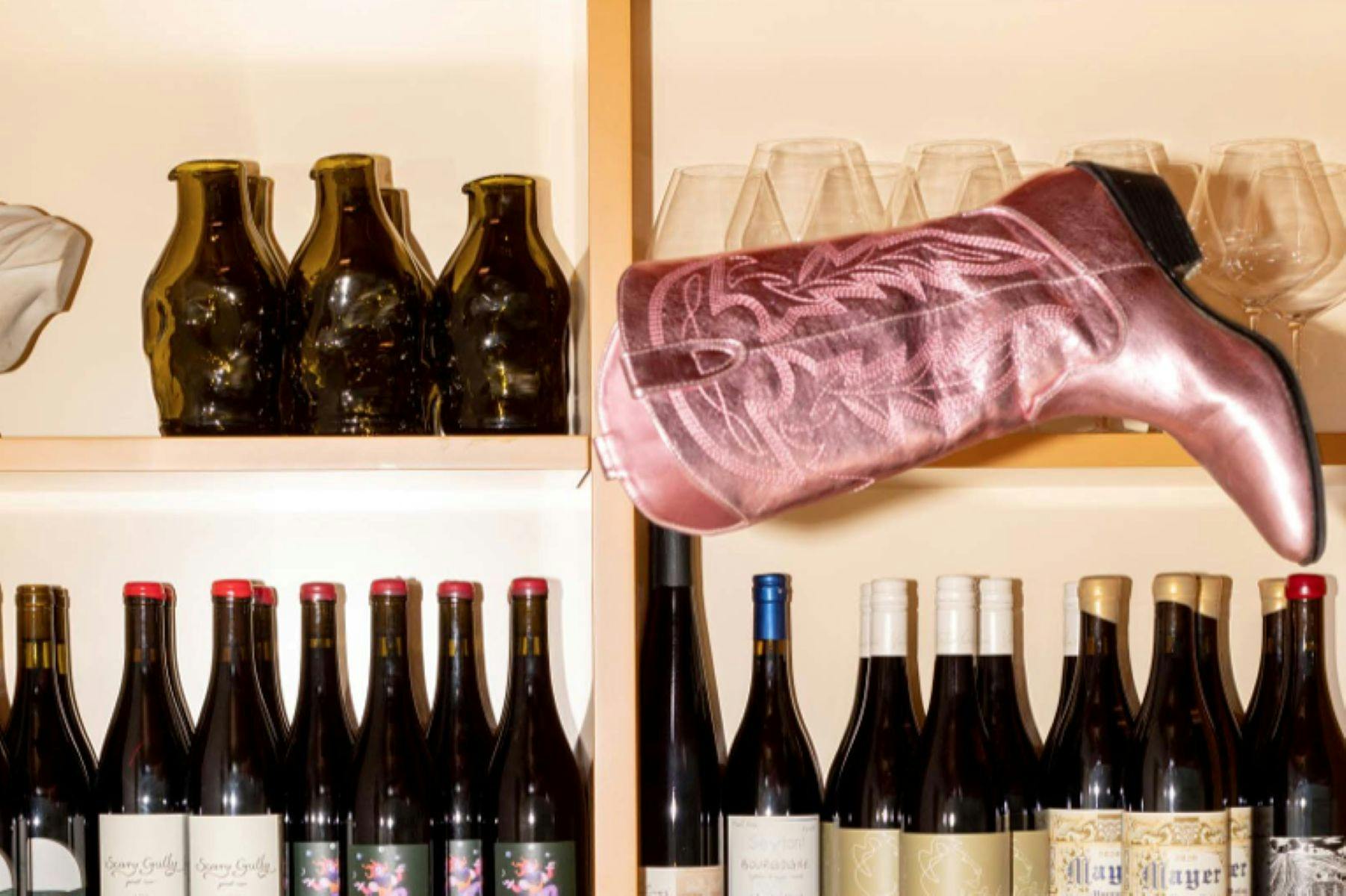 A metallic pink cowboy boot sits at a peculiar angle on a shelf full of wine