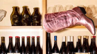 A metallic pink cowboy boot sits at a peculiar angle on a shelf full of wine