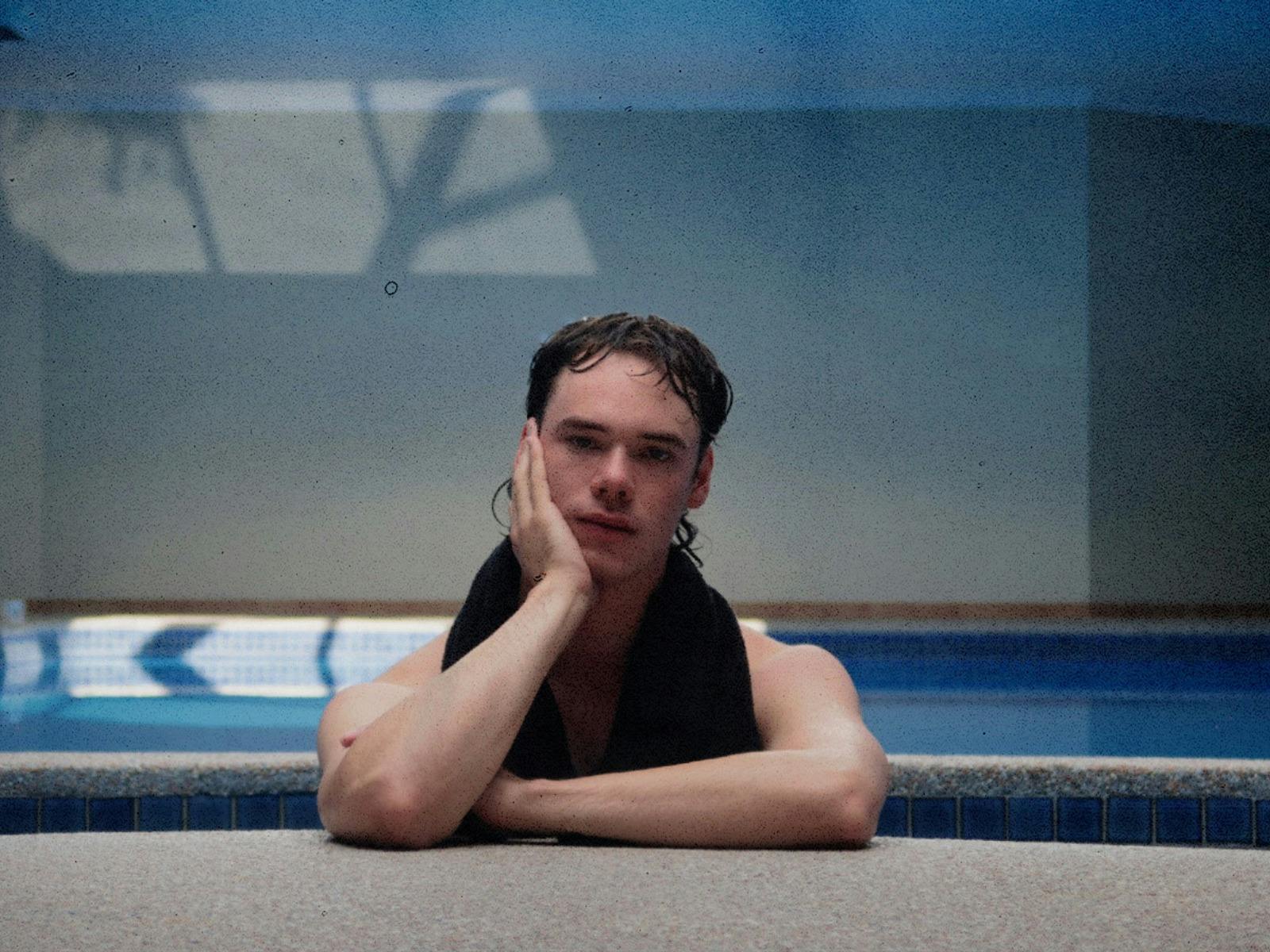 A man leans over the side of a pool and looks directly into the camera.