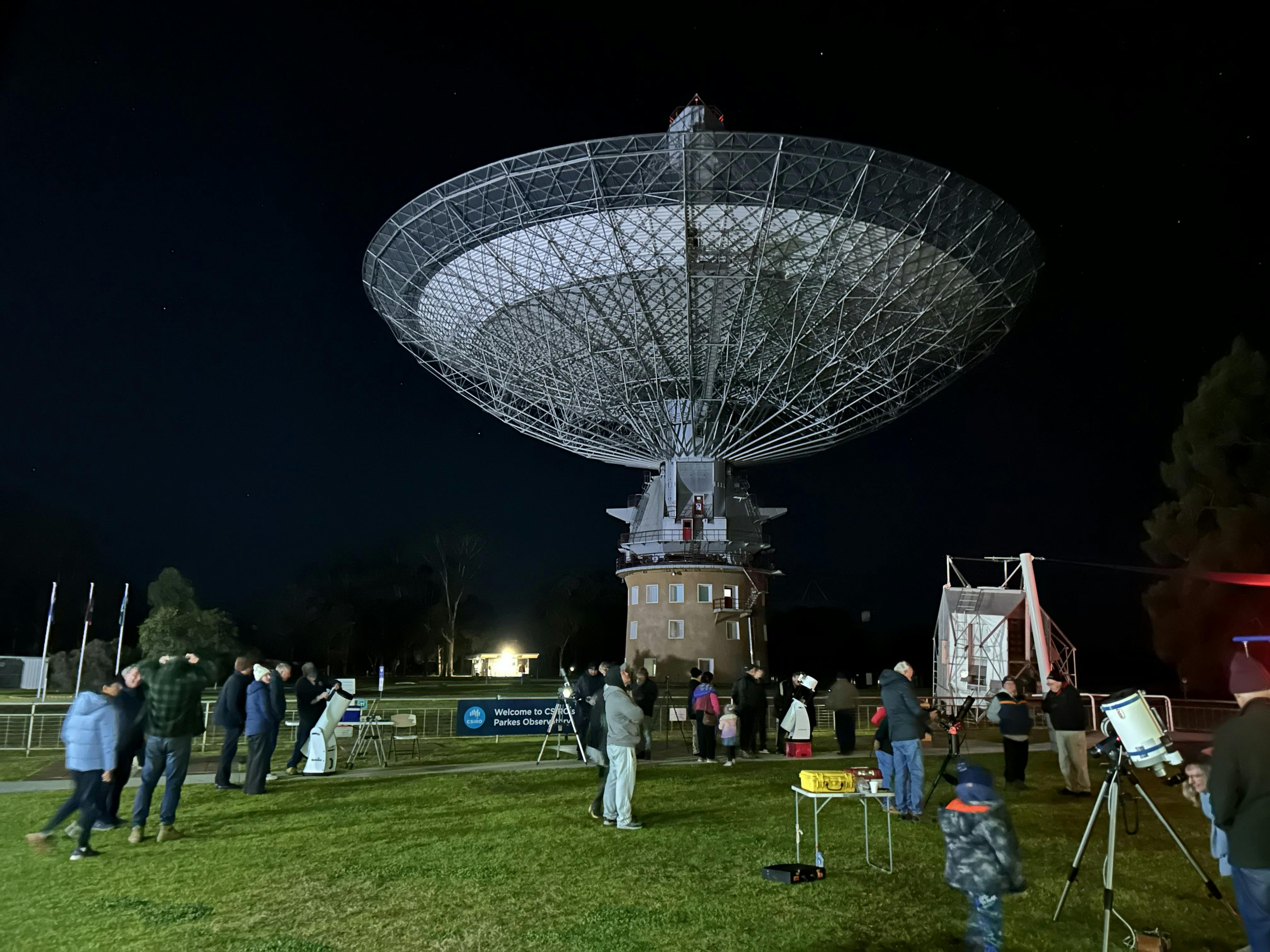 Night view of people looking through optical telescopes with Murriyang in the background