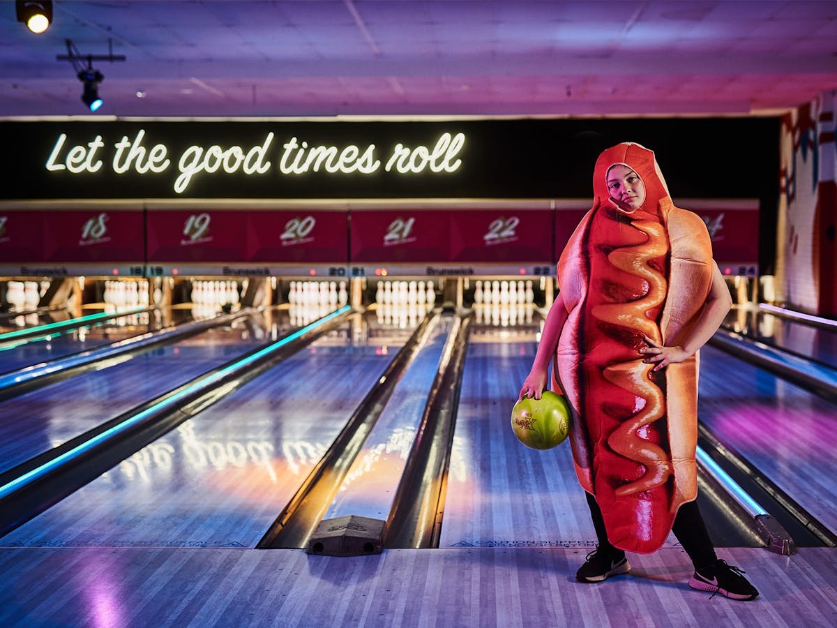 A young person stands in a hotdog costume, holding a bowling ball in front of a bowling alley..