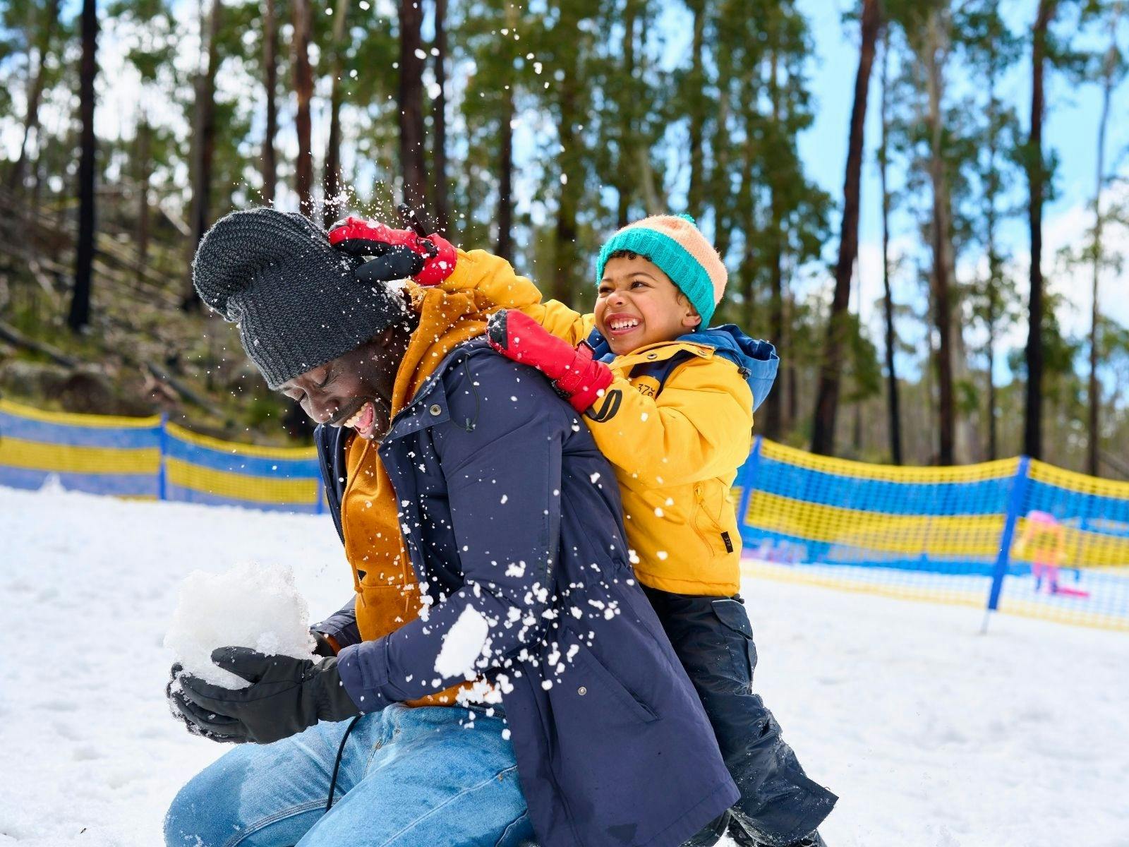 Father kneels in snow as son crushes a snowball on the back of his head, both are laughing