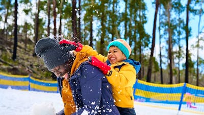 Father kneels in snow as son crushes a snowball on the back of his head, both are laughing