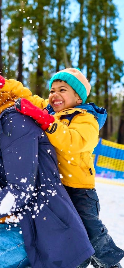Father kneels in snow as son crushes a snowball on the back of his head, both are laughing