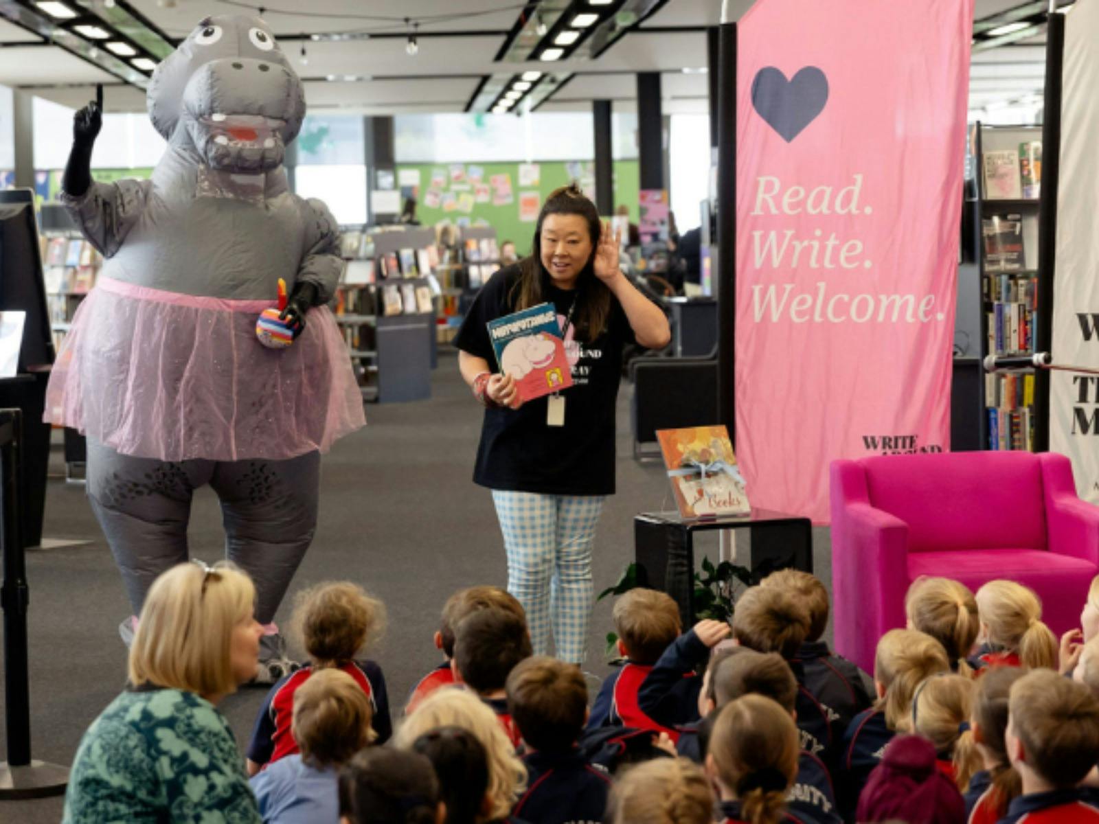 Presenter and a hippo in a pink tutu entertaining seated school children in a library.