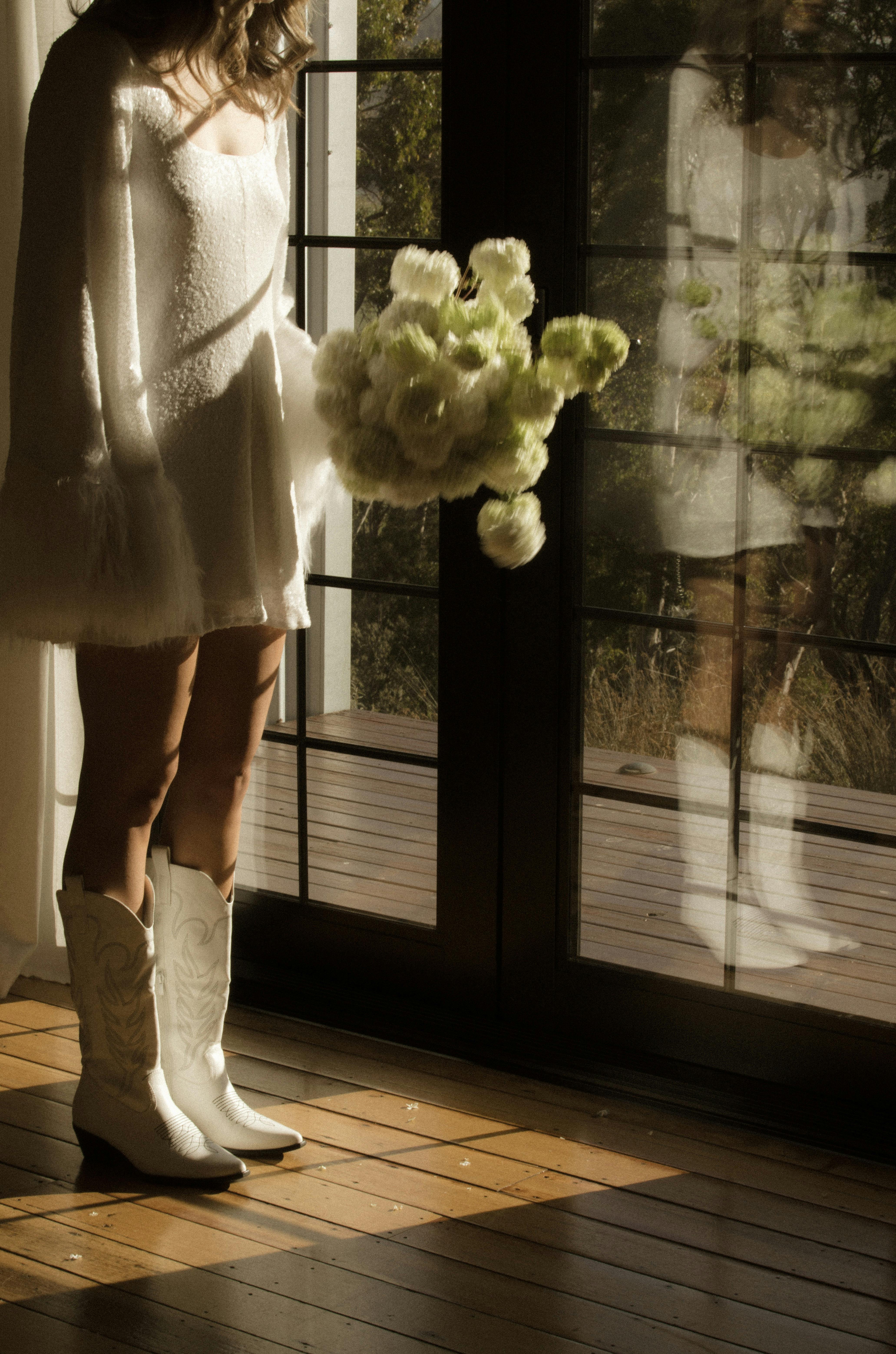 A woman in a short wedding dress holding a bouquet of white flowers stands in front of french doors