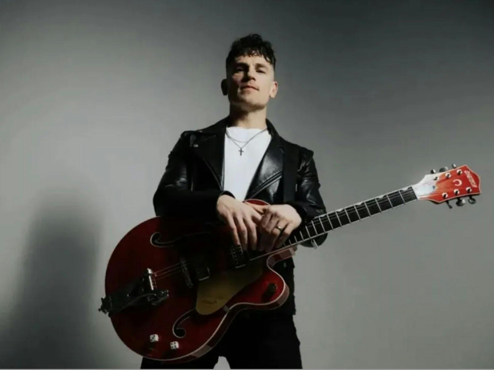 The image is a low-angle, high-contrast portrait of a musician against a neutral grey background. Th