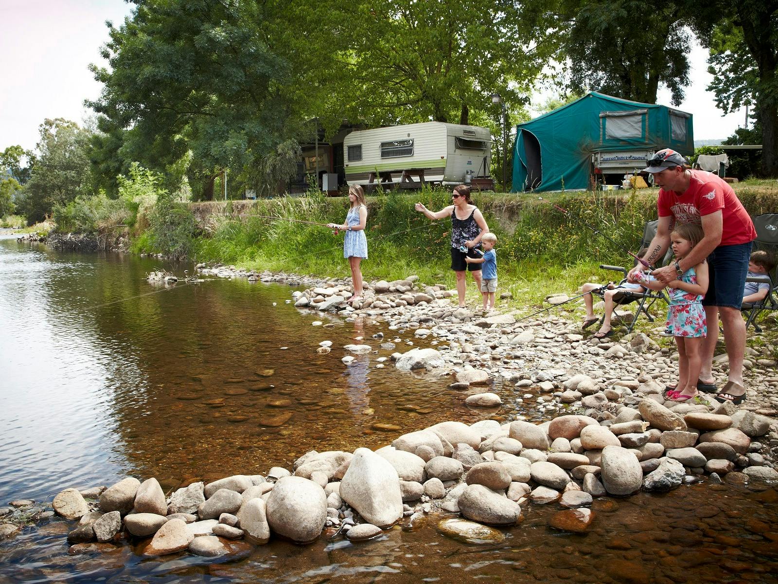 2 adults and 3 kids, fishing off the rocky bank by the river, caravans, tents, sunny day.