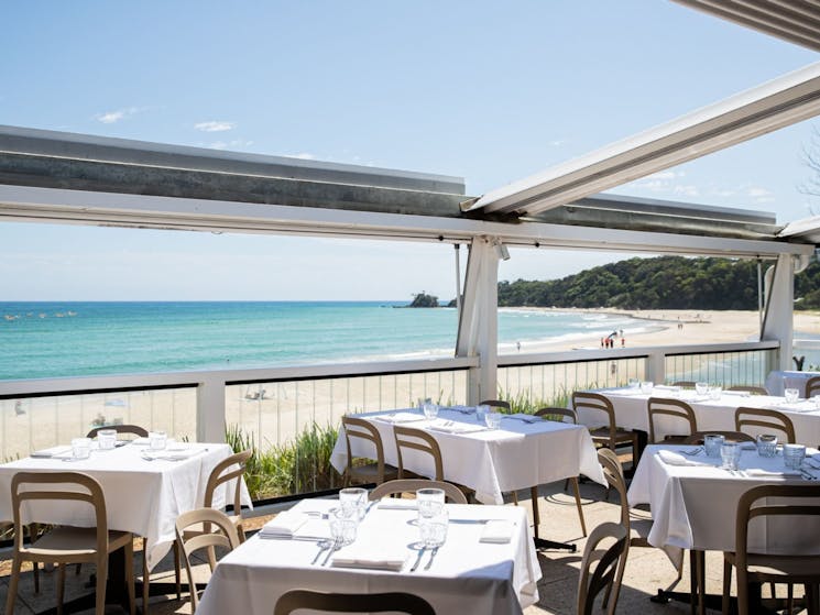 Open air restaurant with views of ocean on a sunny day. Dining tables set with white table cloths.