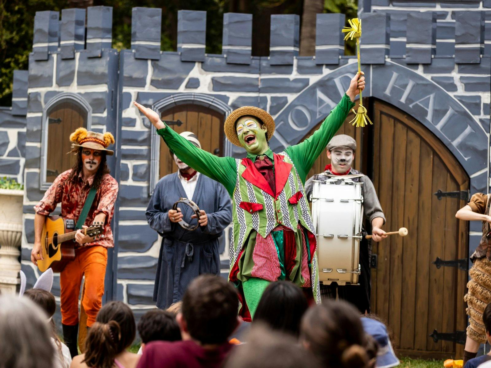 Four performers dressed as animals play instruments and sing in front of a castle set piece.