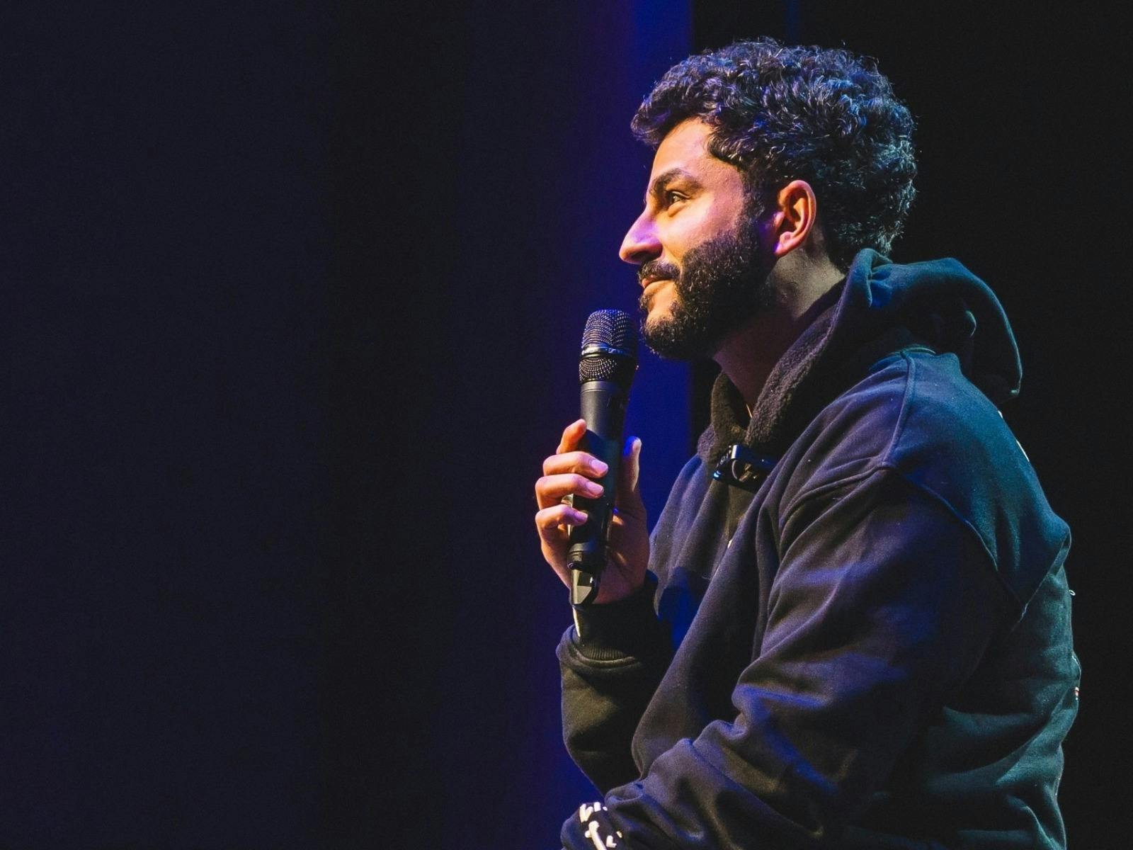 A smiling man with a beard holds a microphone while speaking on stage against a dark backdrop.