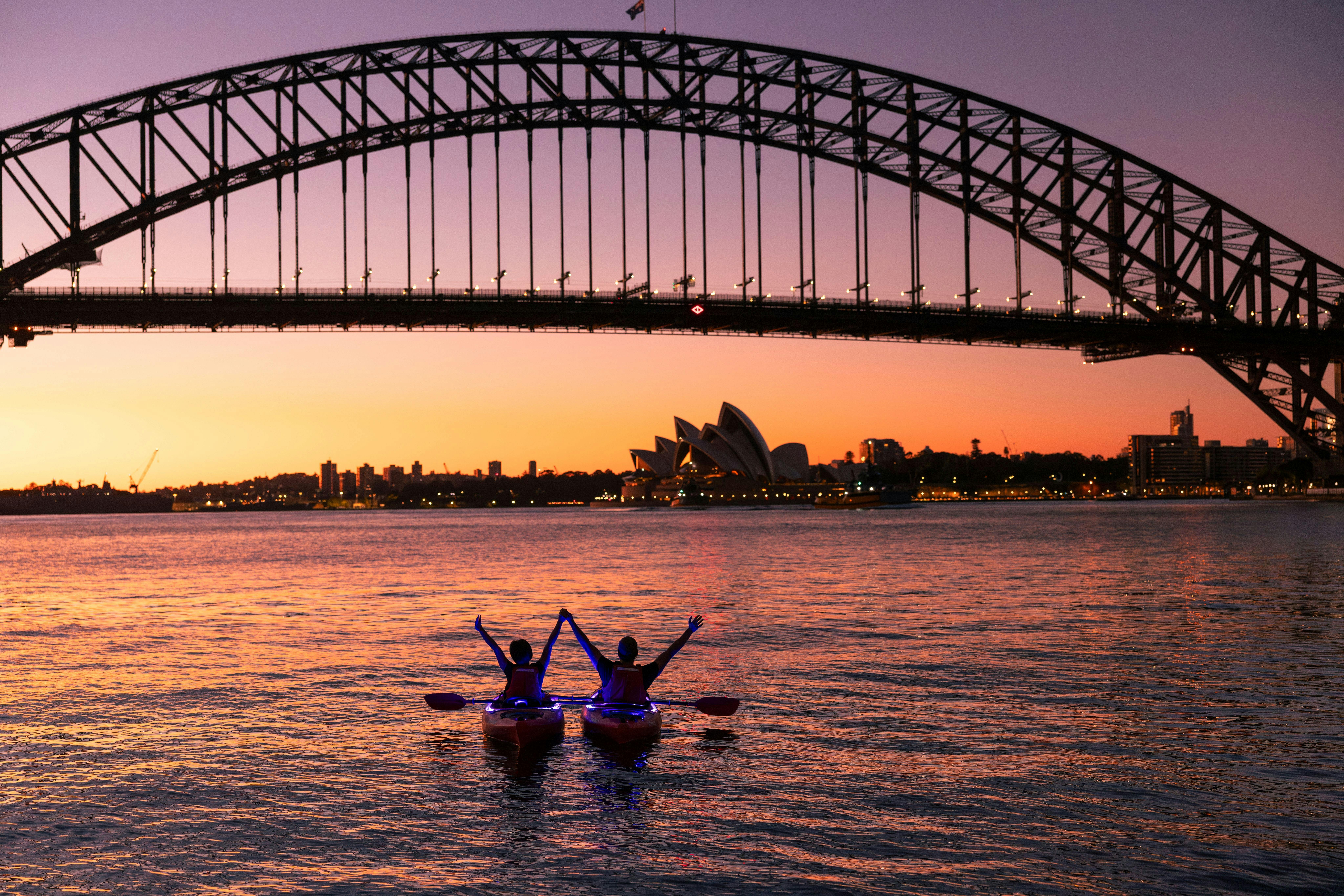 Two people paddling holding hands at sunrise with the Sydney Harbour bridge and golden glow