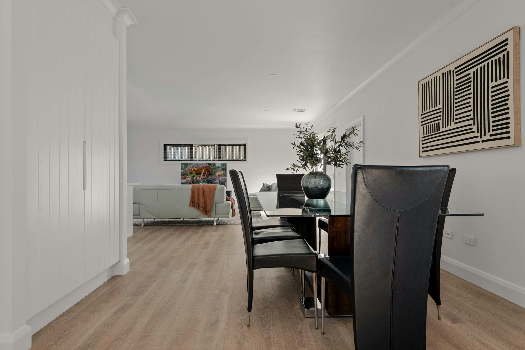 Glass dining table with leather black chairs, couch in the background. White walls and wooden floors