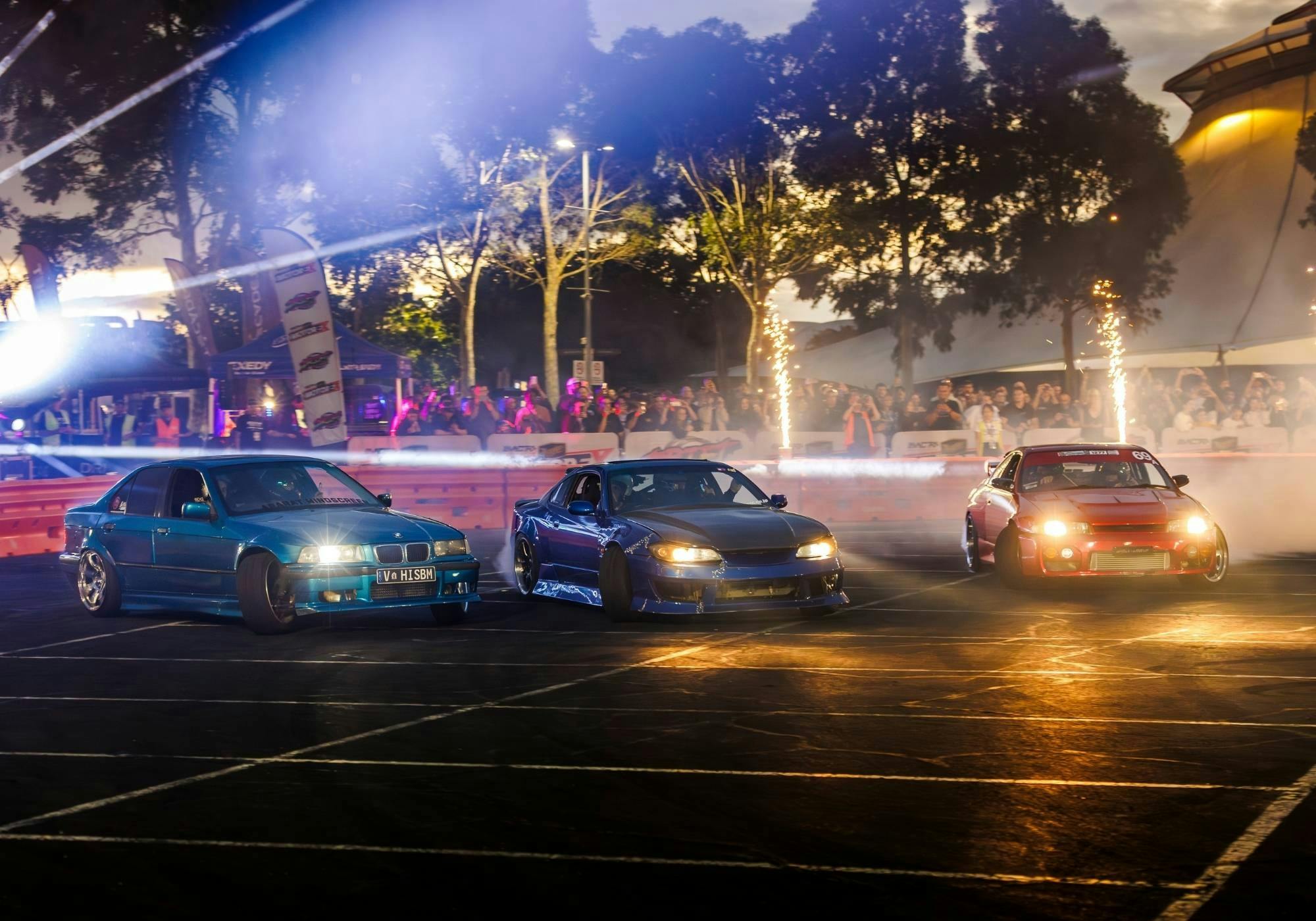 Three modified drift cars sliding across the course at night beneath lights and pyrotechnics.