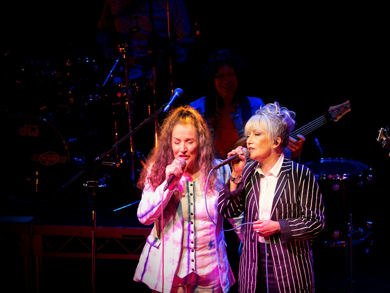 Two women - Wendy Matthews and Grace Knight singing on microphones on a darkened stage.