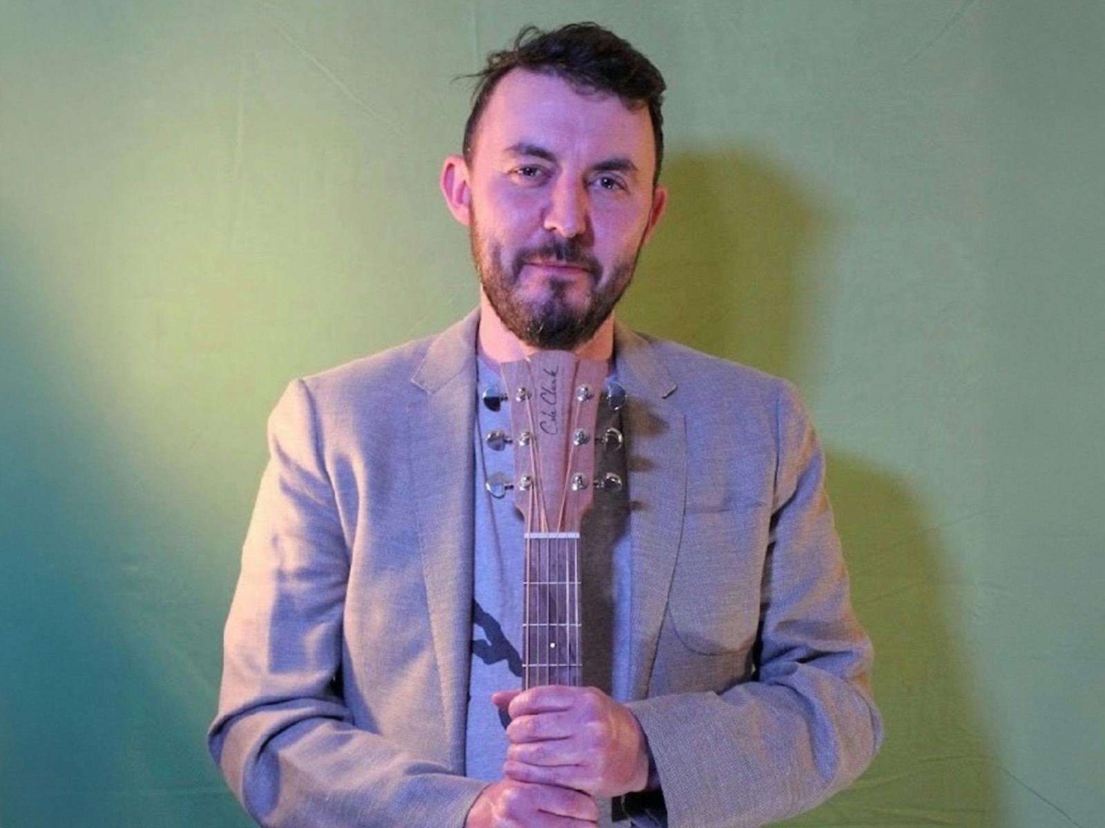 Bearded man wearing a grey suit holding a guitar