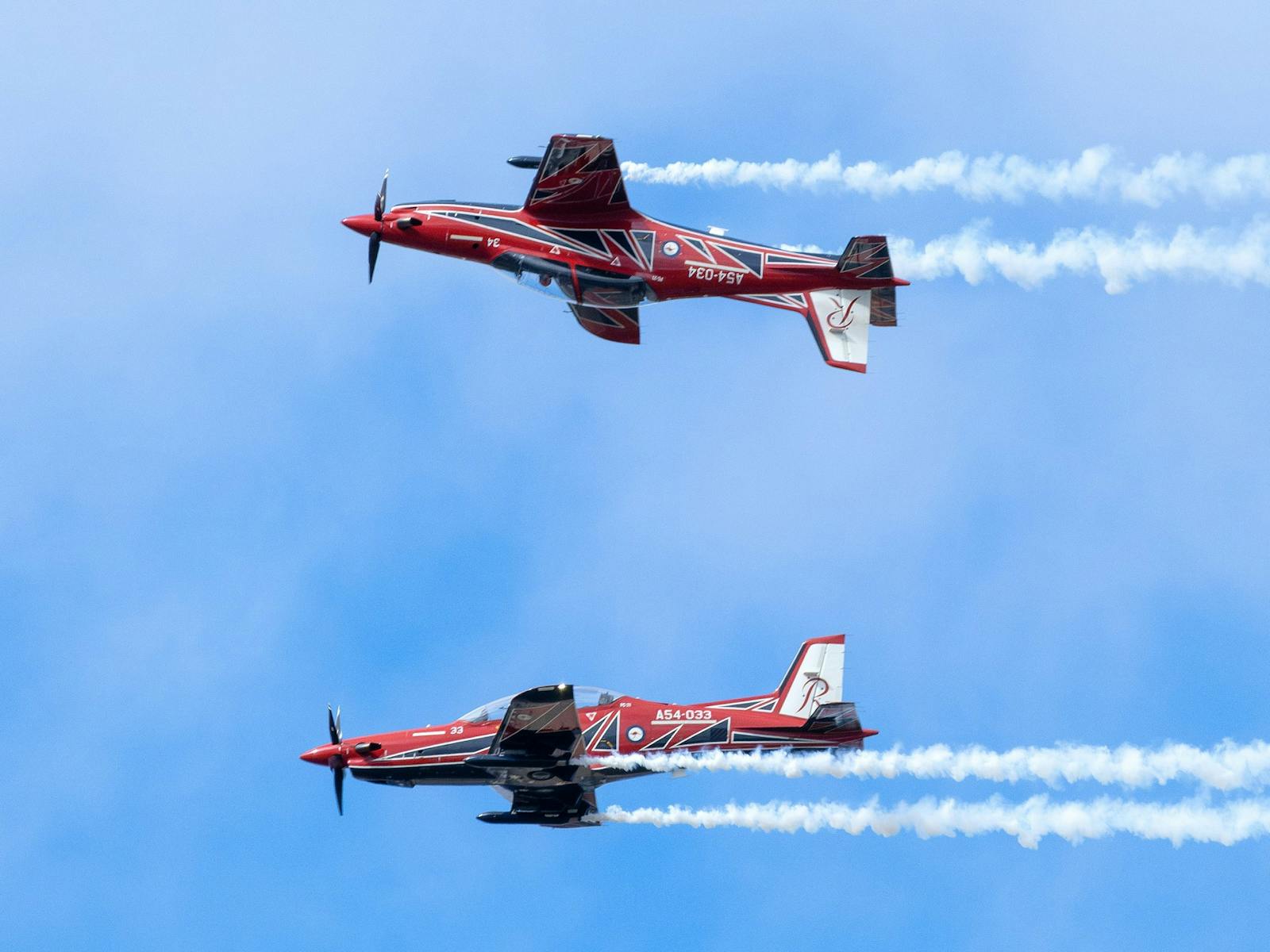 Two red-and-white Pilatus PC-21 aircraft fly overhead with display smoke streaming behind