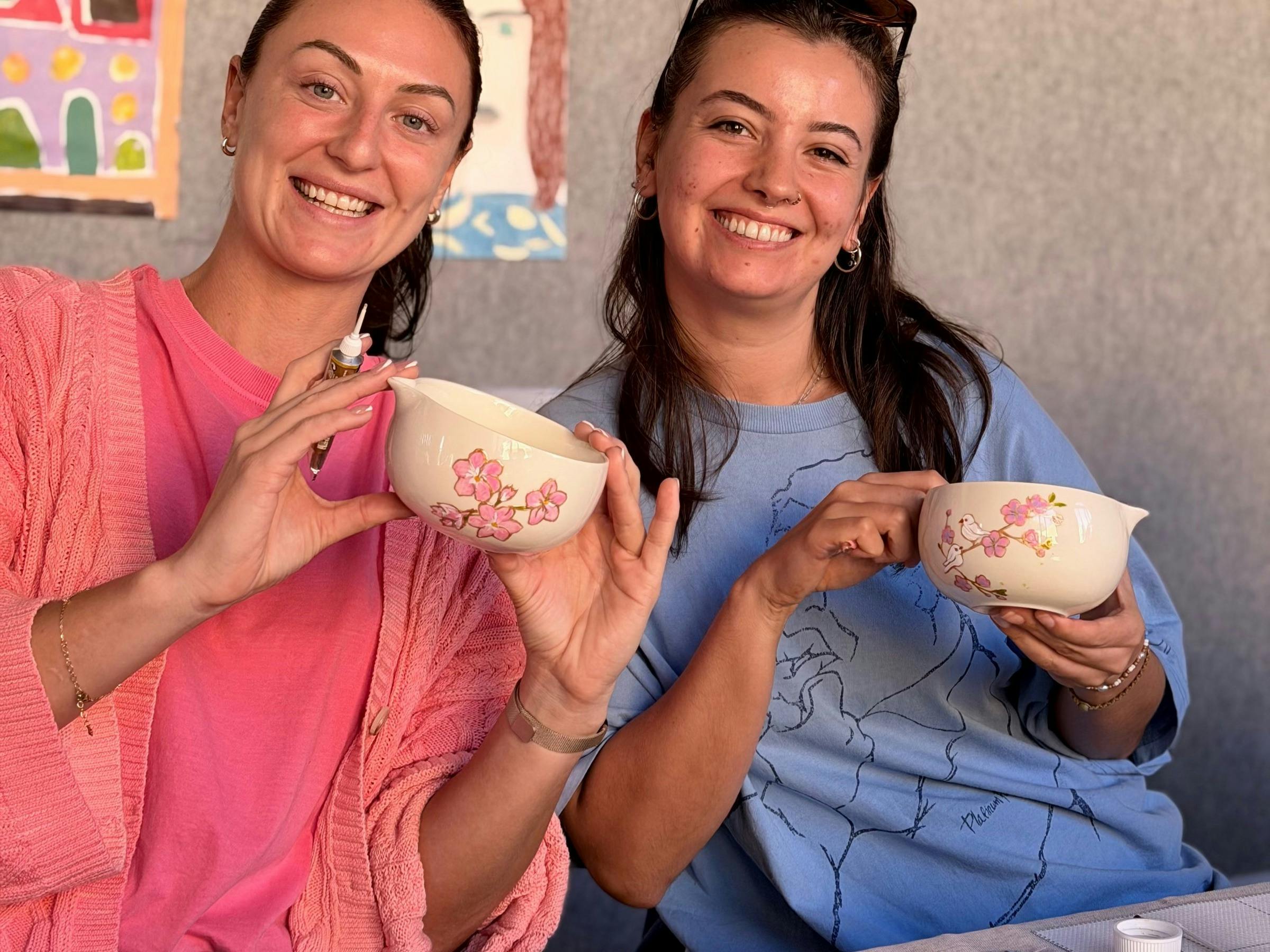 Two smiling women holding ceramic matcha bowls they painted with pink cherry blossoms