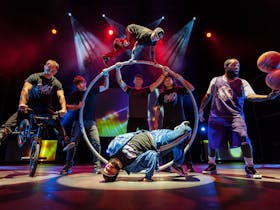 7 men on stage performing various acrobatic and sports themed tricks.