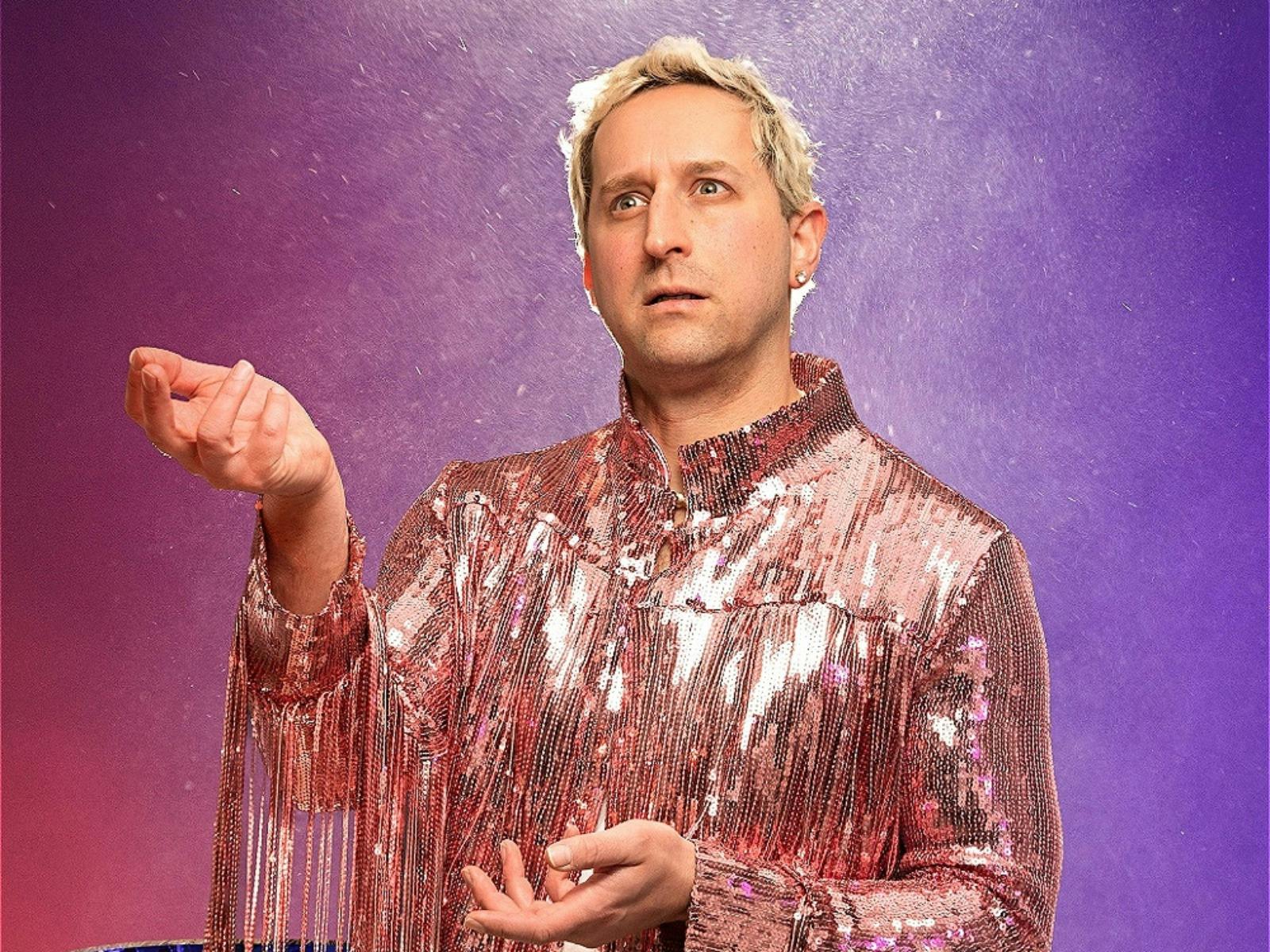 A blonde man in a pink sequined top stares blankly against a vibrant purple-red background.