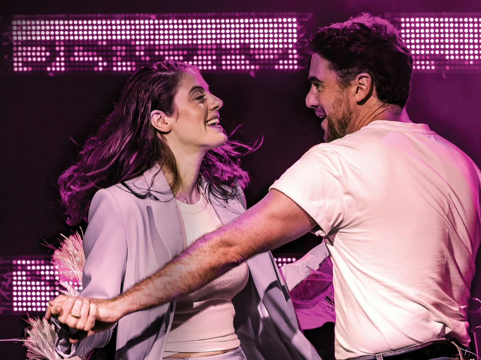 A young woman and a man smile at each other while dancing on stage, illuminated by bright pink LED