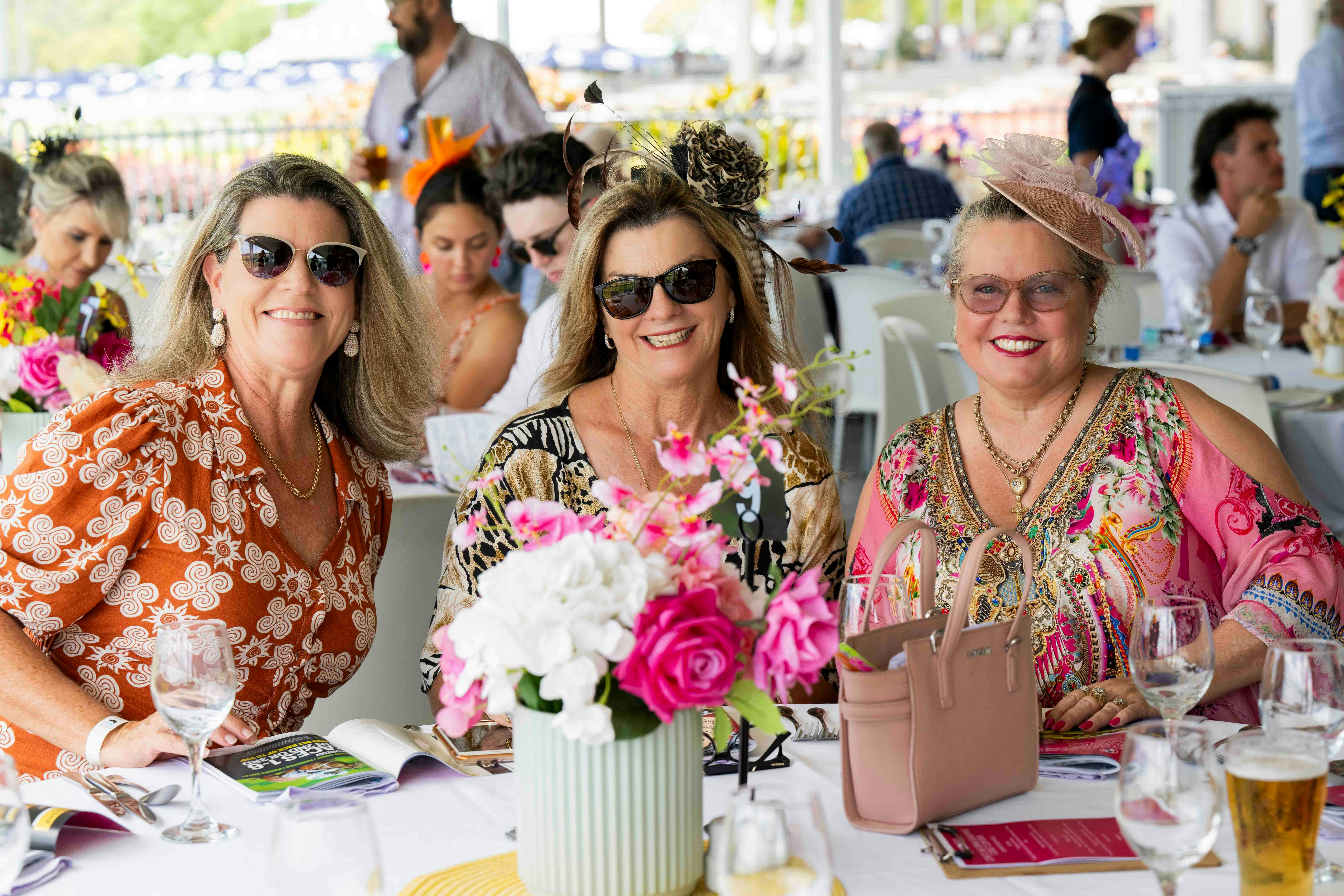 Three ladies siting at a table with a floral arrangement in the middle smiling