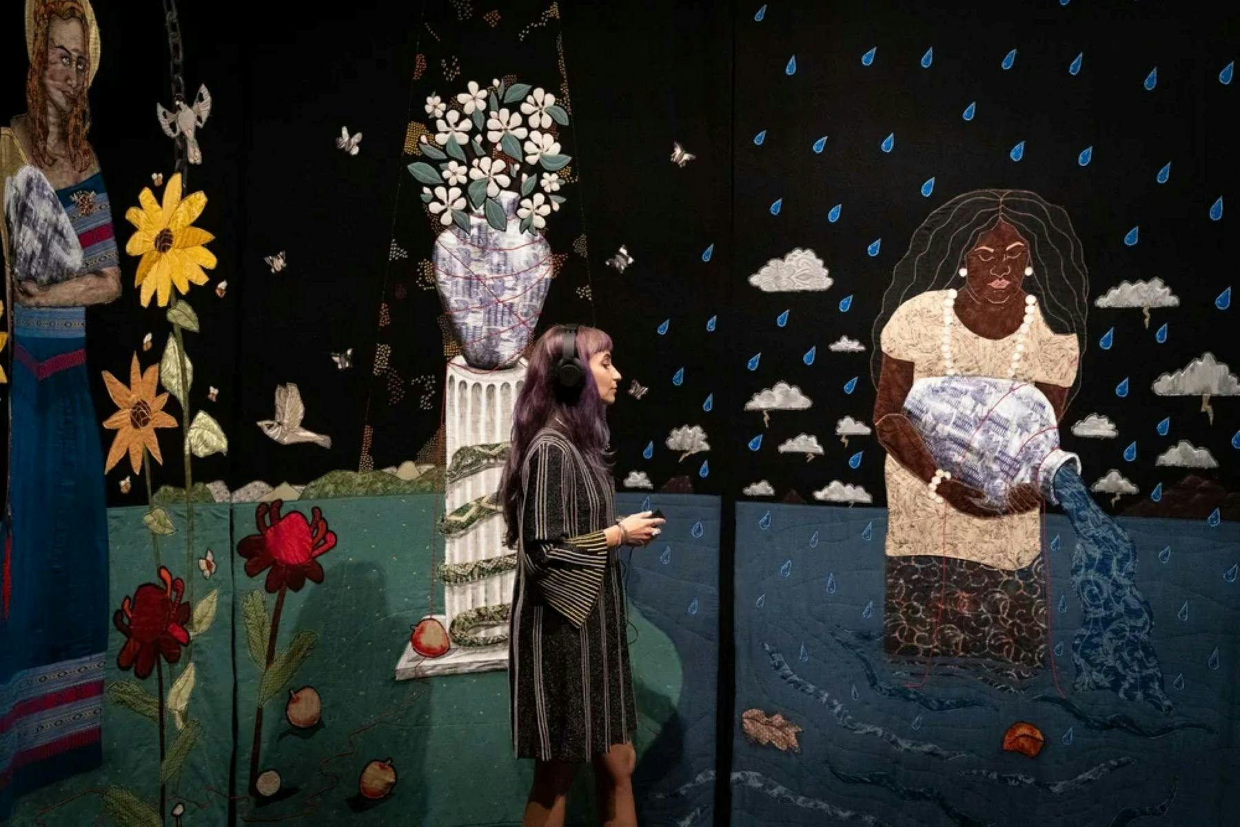 A woman stands in front of a large tapestry artworks with flowers and a woman with a water vessel