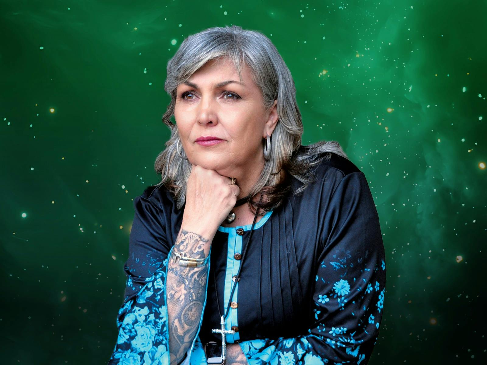 A white woman with grey hair, wearing a blue flowy shirt. Her chin is resting on her hand.