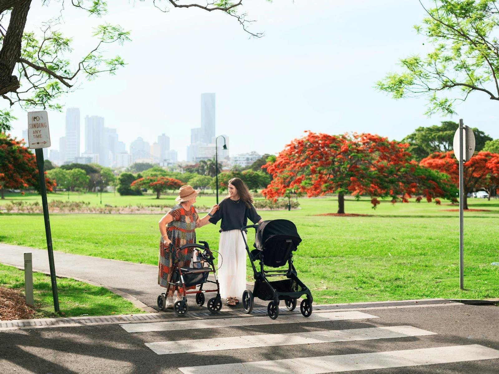 A younger woman helping an elderly woman with a walker cross a pedestrian crossing in a park, with a A younger woman helping an elderly woman with a walker cross a pedestrian crossing in a park, with a