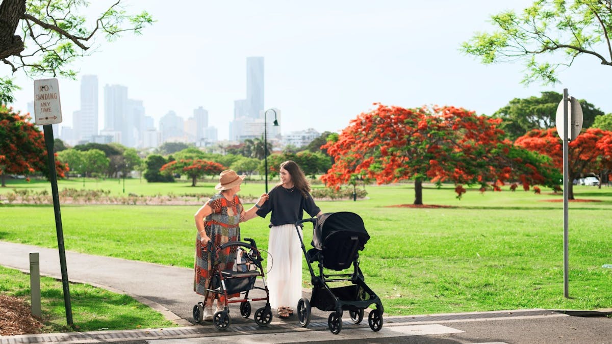 A younger woman helping an elderly woman with a walker cross a pedestrian crossing in a park, with a