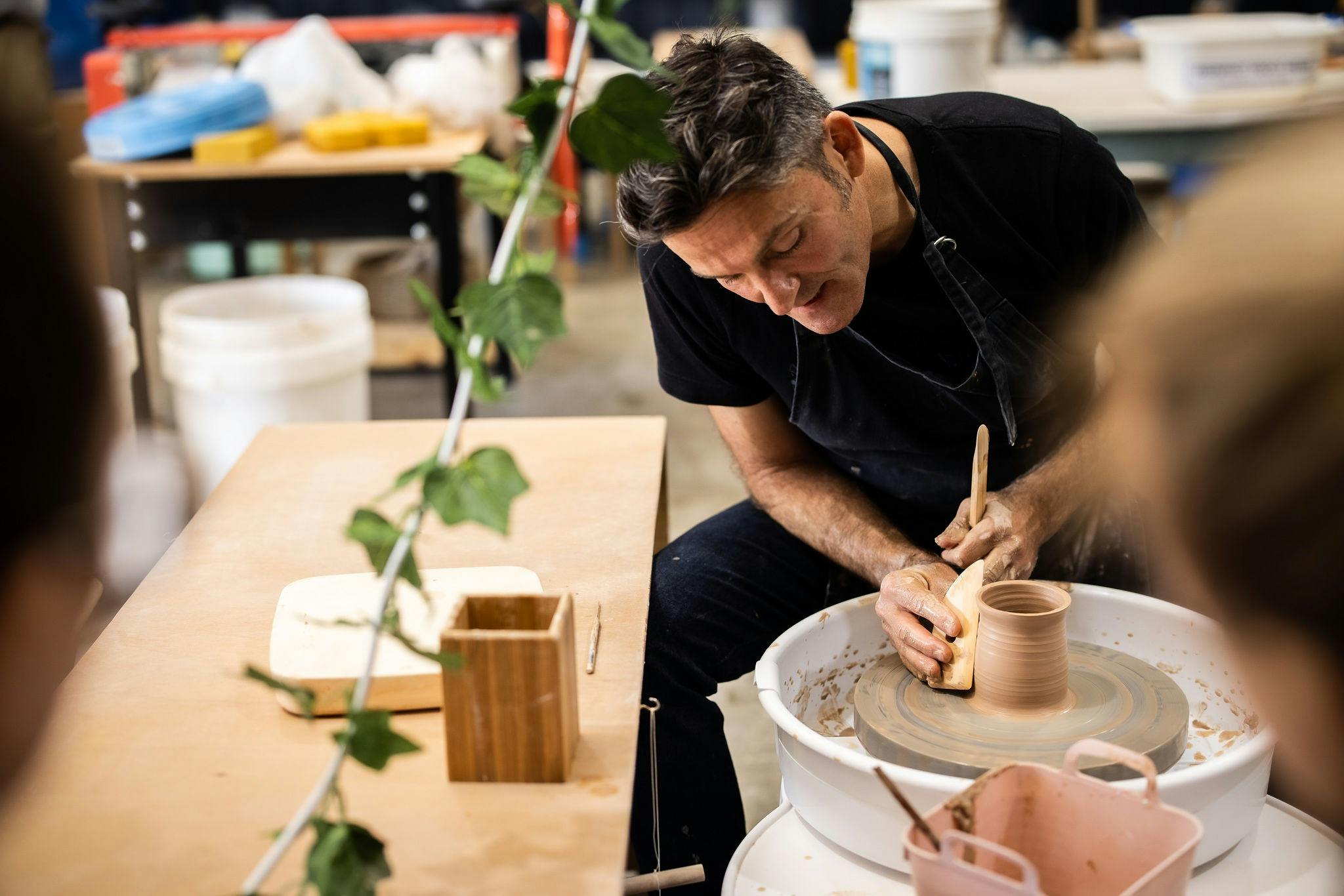 Picture shows a male seated at a pottery wheel with a pottery tool in his hand working with the clay