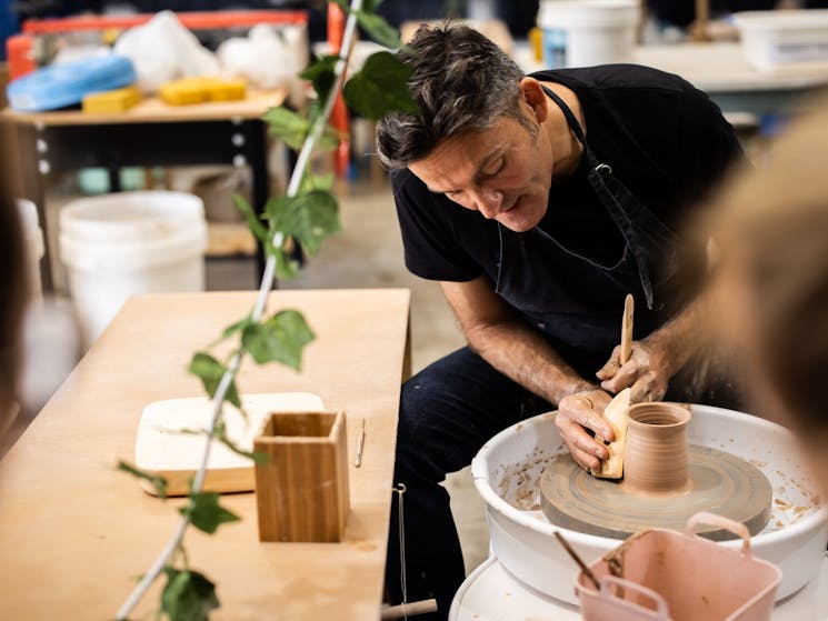 Picture shows a male seated at a pottery wheel with a pottery tool in his hand working with the clay