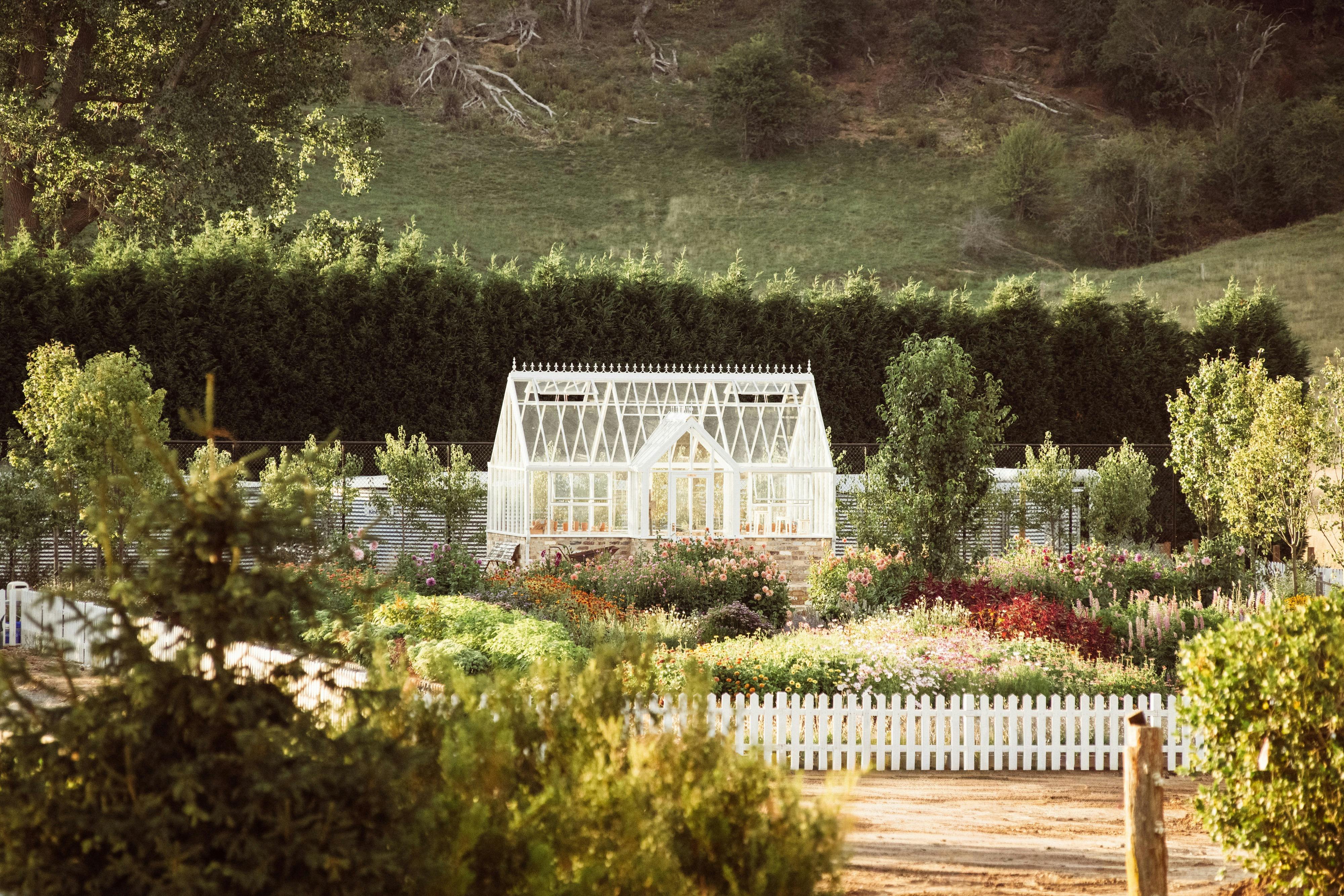 A white glass greenhouse surrounded by a colorful flower garden and trees.