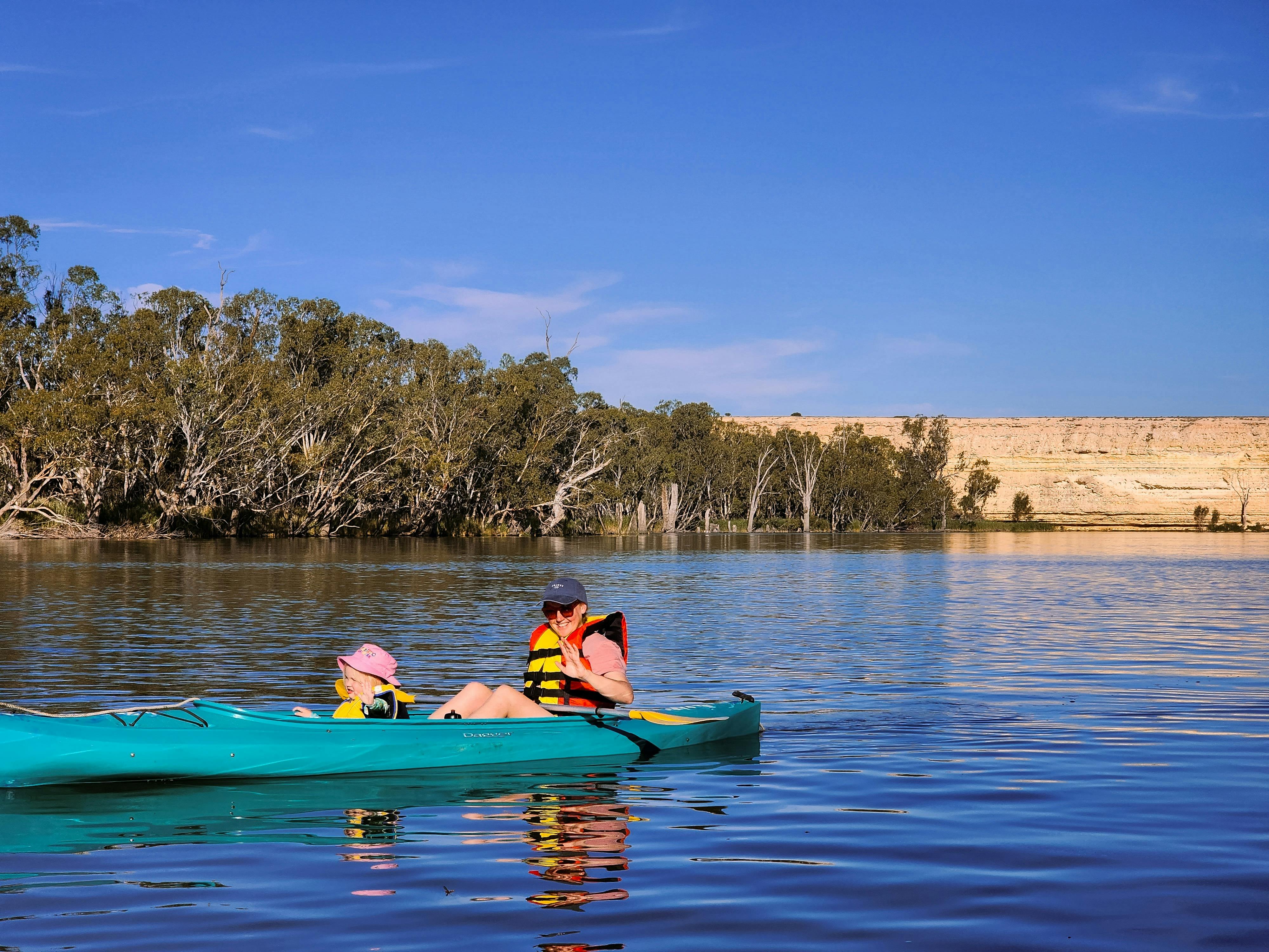Adult and child kayaking on the Murray River with river red gums and golden limestone cliffs.