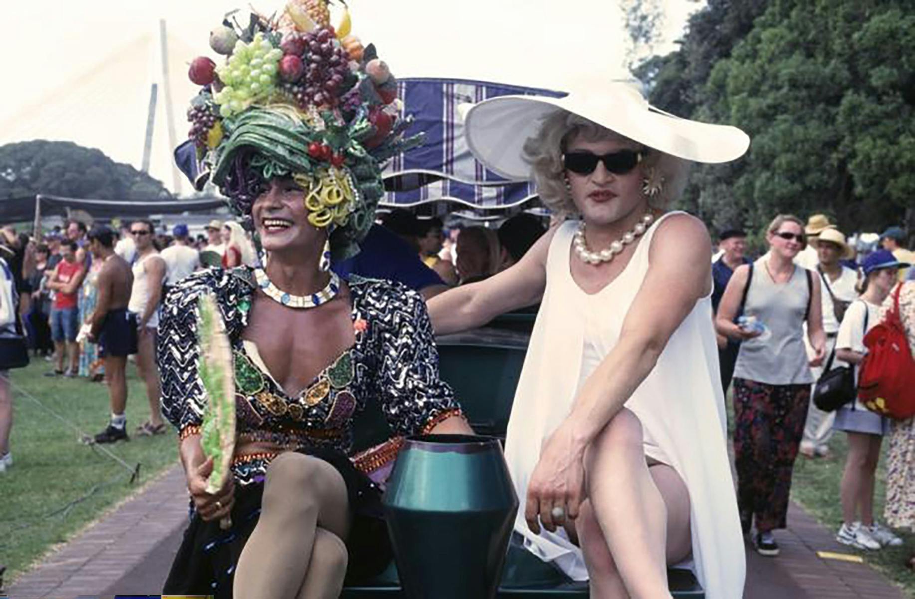 One man dressed in a vibrant dress with a hat with fruits on it and a man in a white dress and hat