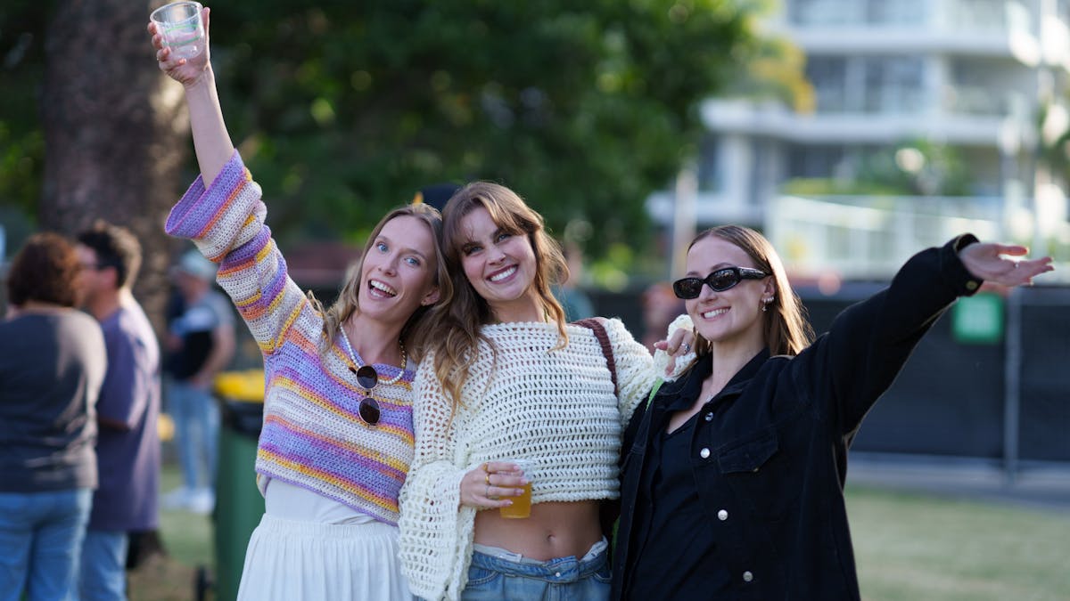 3 girls face the camera smiling in an embrace while holding a beer to the sky