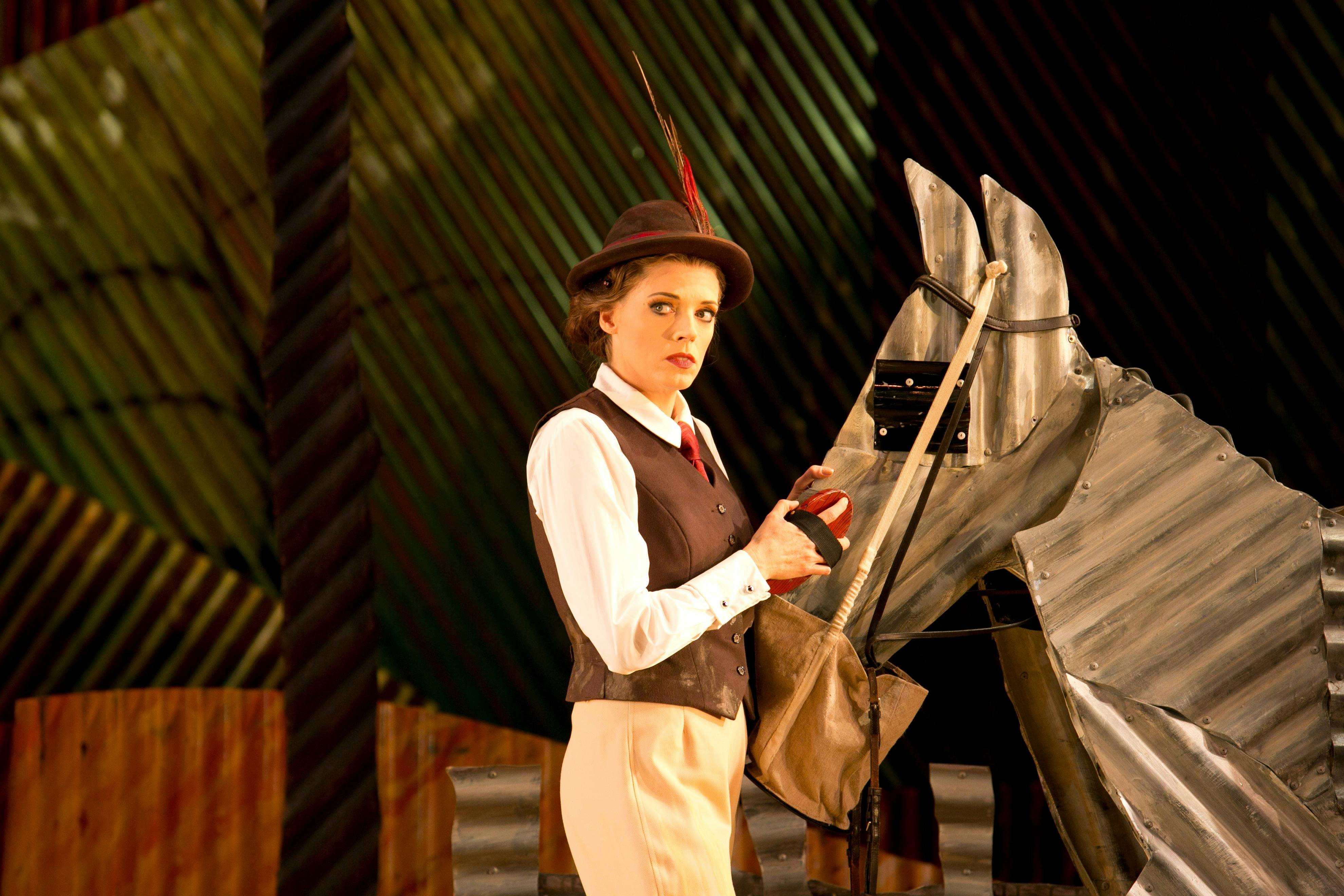A woman in period riding gear stands beside a horse made of corrugated iron