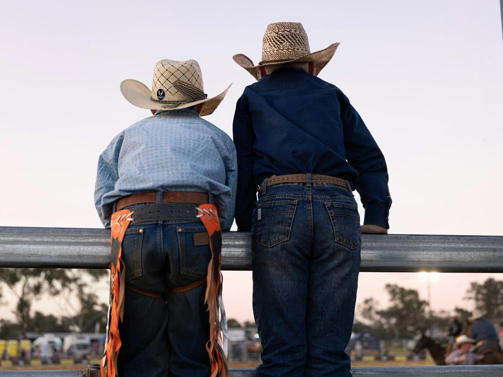 two boys standing on a fence watching the horses, both have jeans and cowboy hats on