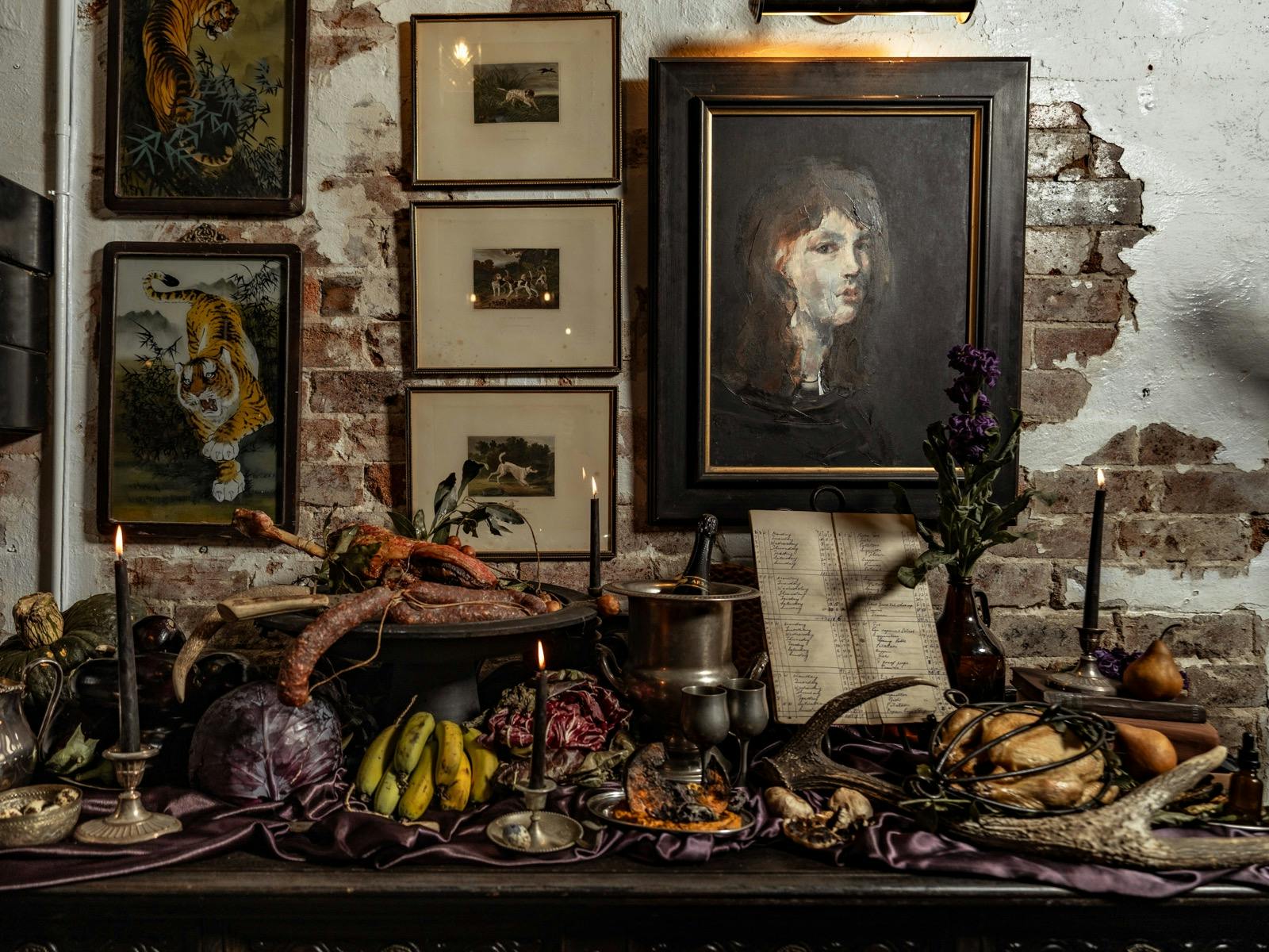A moody Earth Walker still life with fire-cooked meats, seasonal produce, candles, vintage ware, and