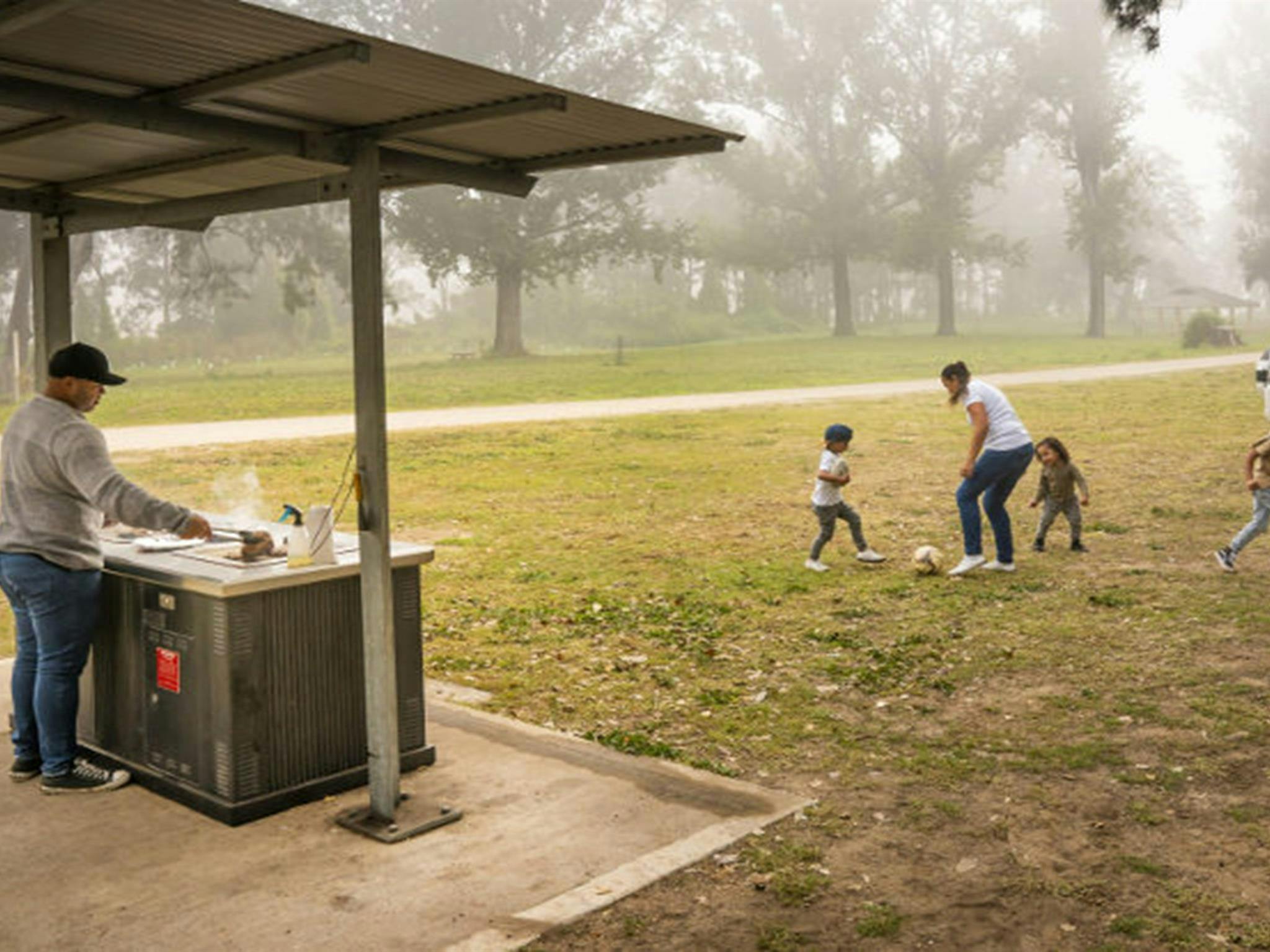 A man cooks up a barbecue lunch as a woman and three kids play soccer in the background. Cattai Farm