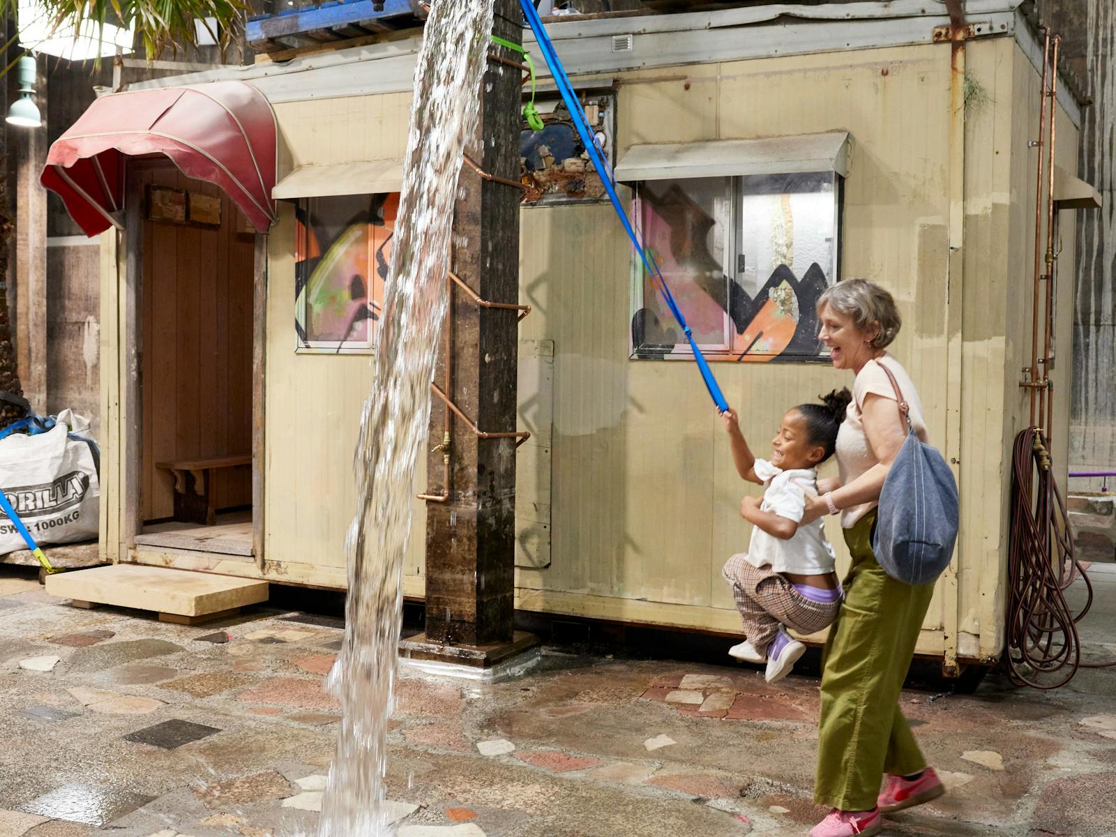 A child and their mother pull a rope that empties a bucket of water in an art gallery space