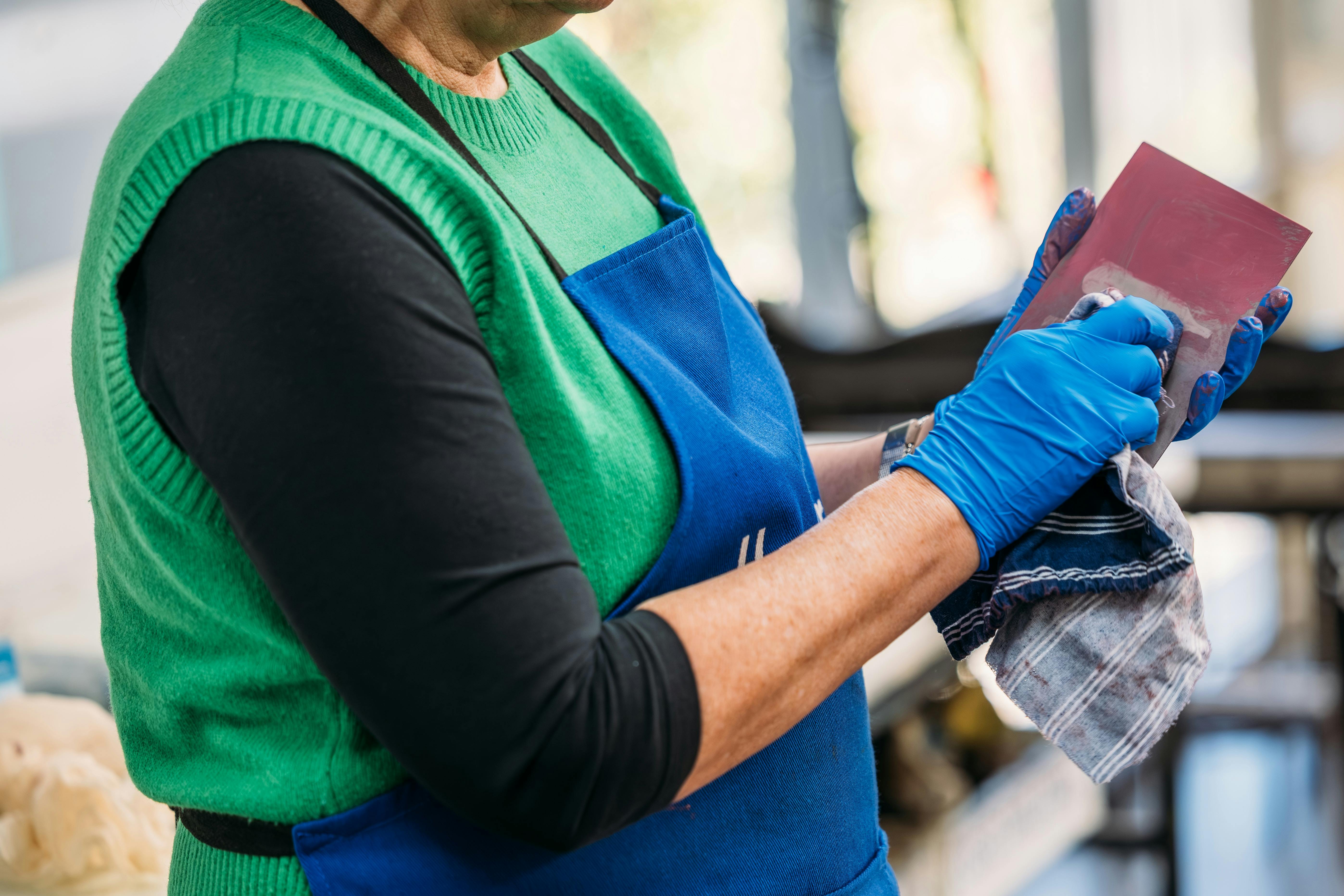 A person wearing a blue apron and blue rubber gloves wipes ink from a small printing plate.