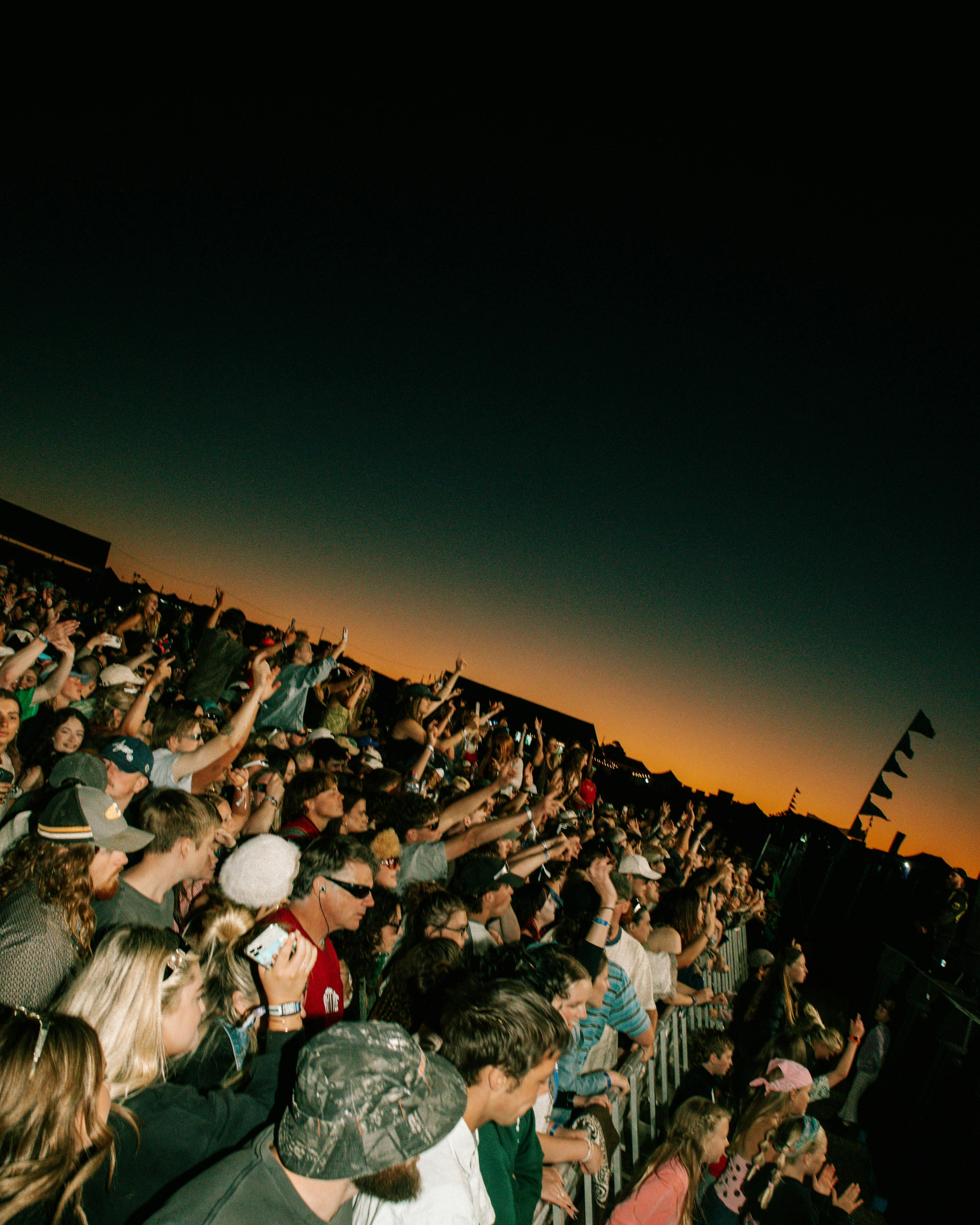 Crowd cheering with raised hands beneath an orange sunset sky at Good Gumnuts Festival.