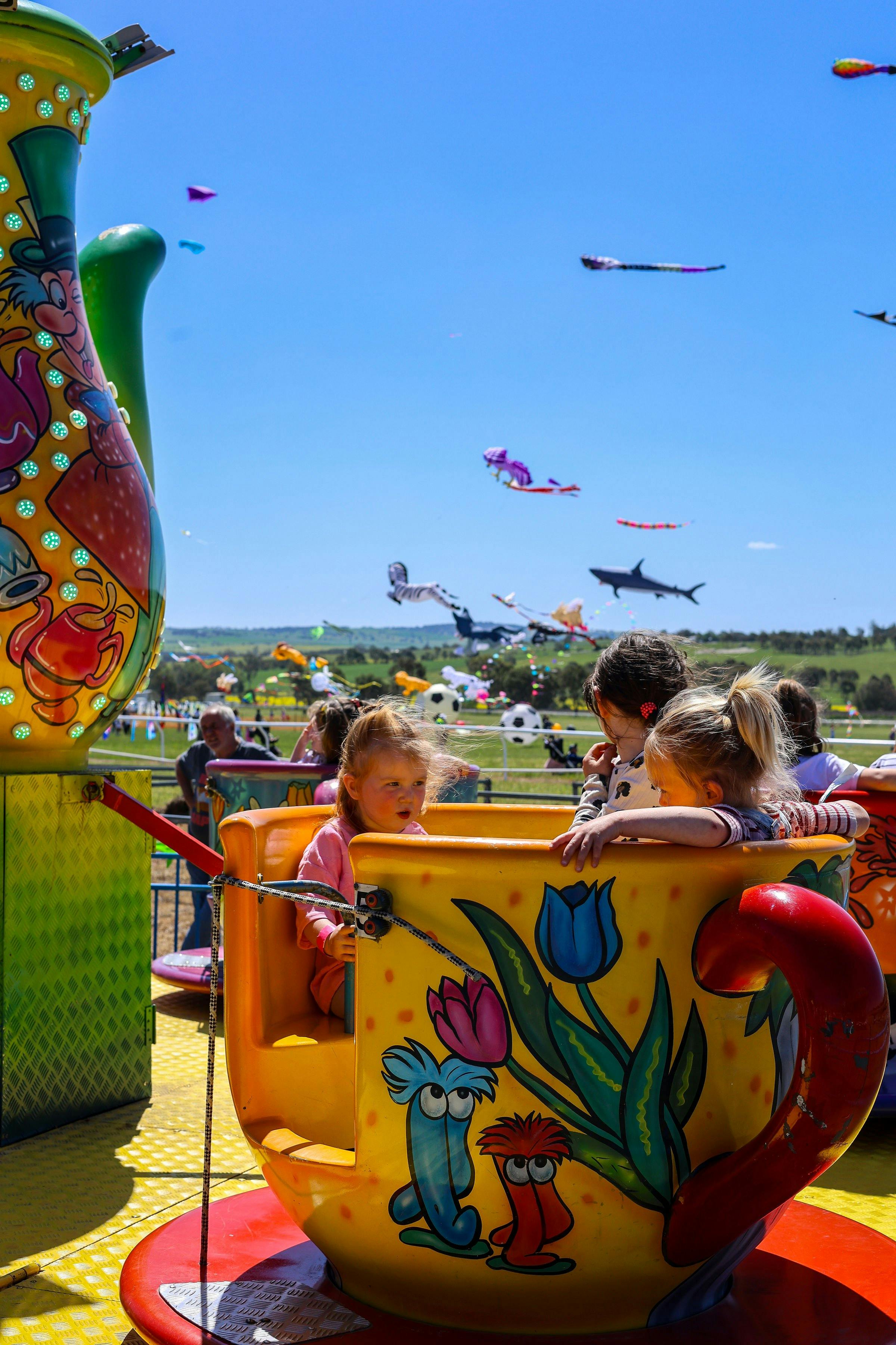 Three children in a teacup ride chatting away while colourful kites soar in the sky.