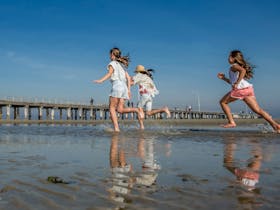 Children running at Altona Beach with Altona pier in the background.