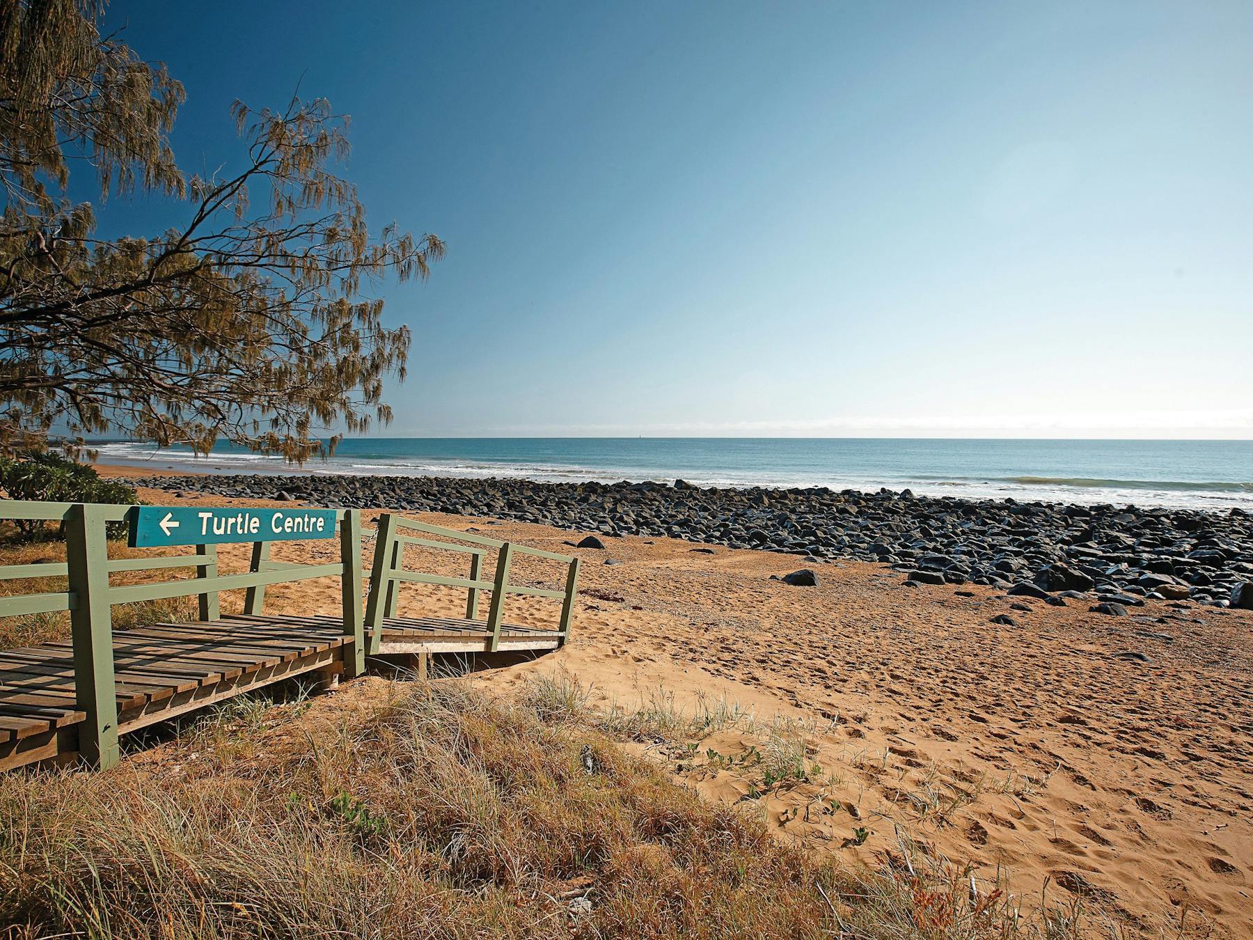 View of Mon Repos sandy beach, volcanic rock and blue ocean beyond with a wooden walkway to the left