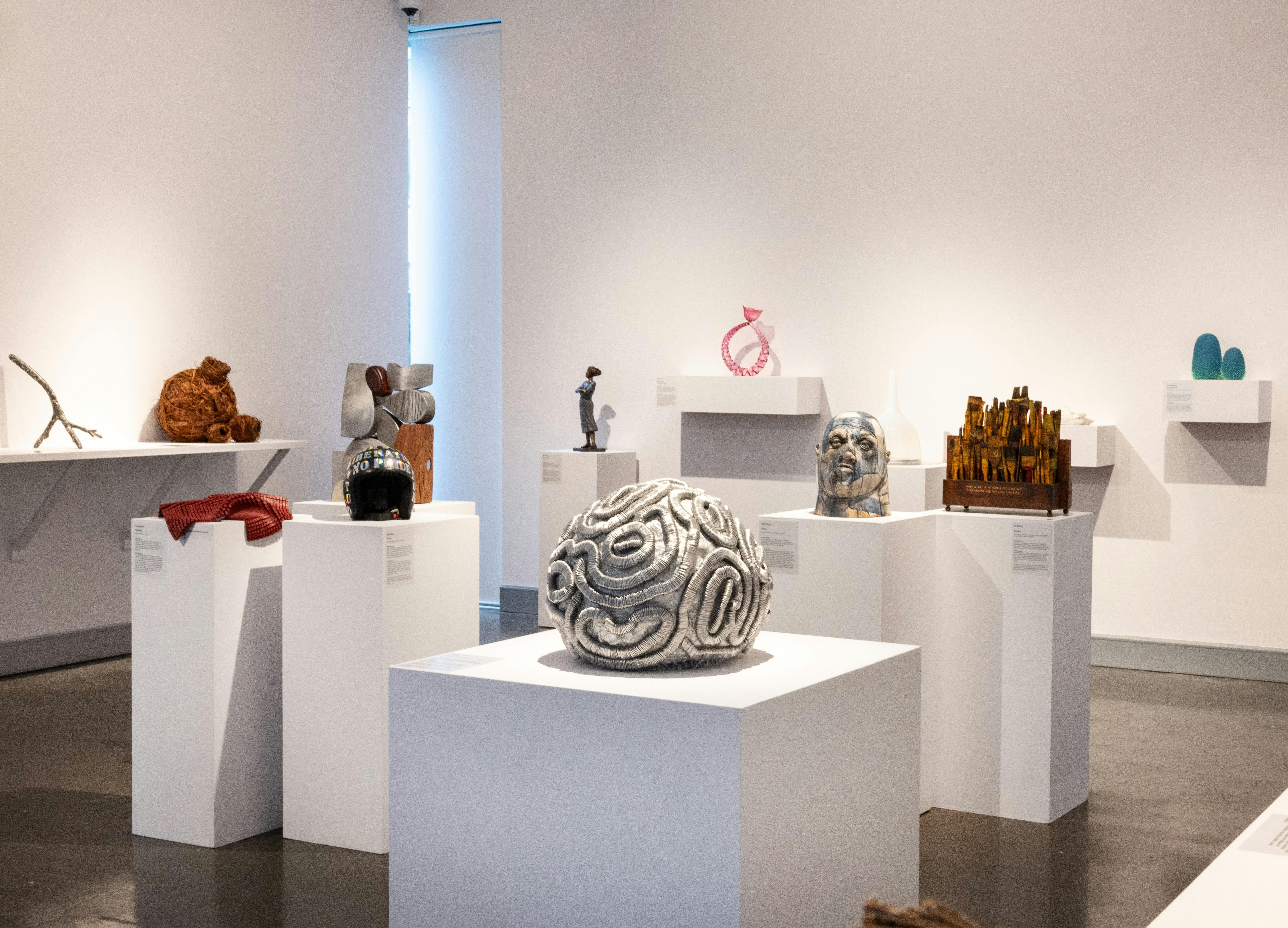 Installation image of sculptures of various mediums displayed on white plinths and wall shelving