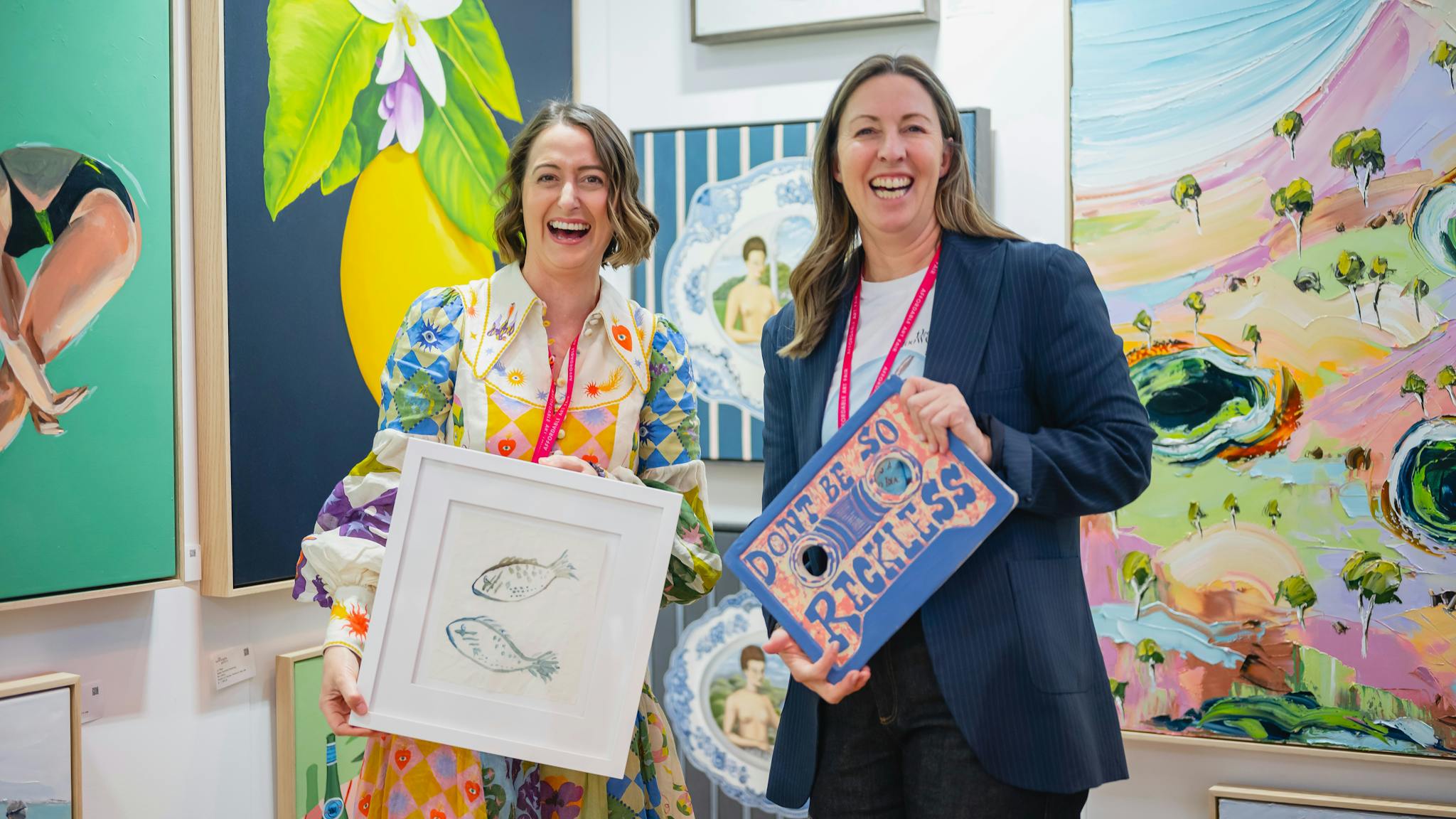 Two friendly gallerists at their Affordable Art Fair stand holding an artwork each