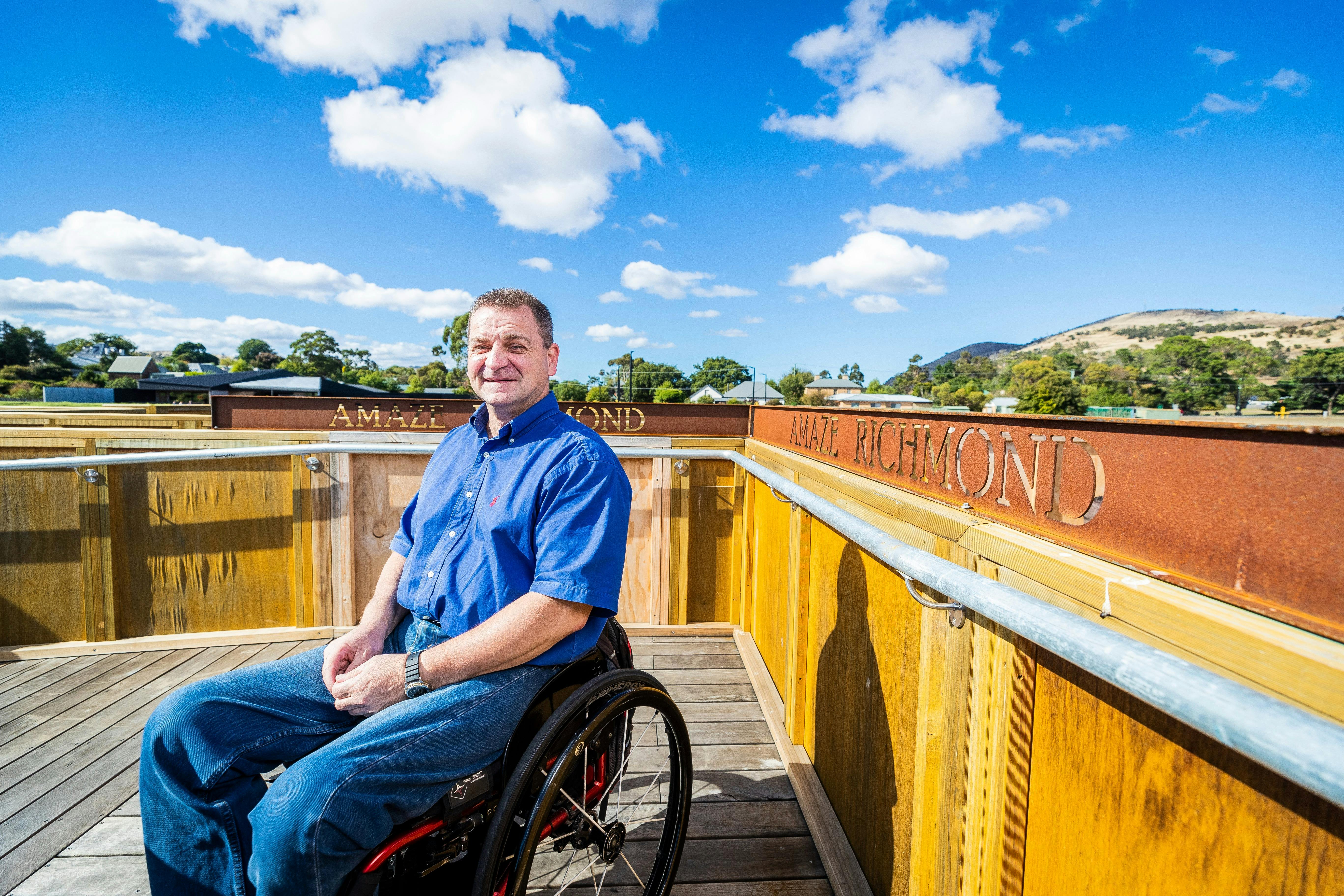Smiling man wearing a blue shirt and jeans, sitting in a wheelchair in front of a metal sign.