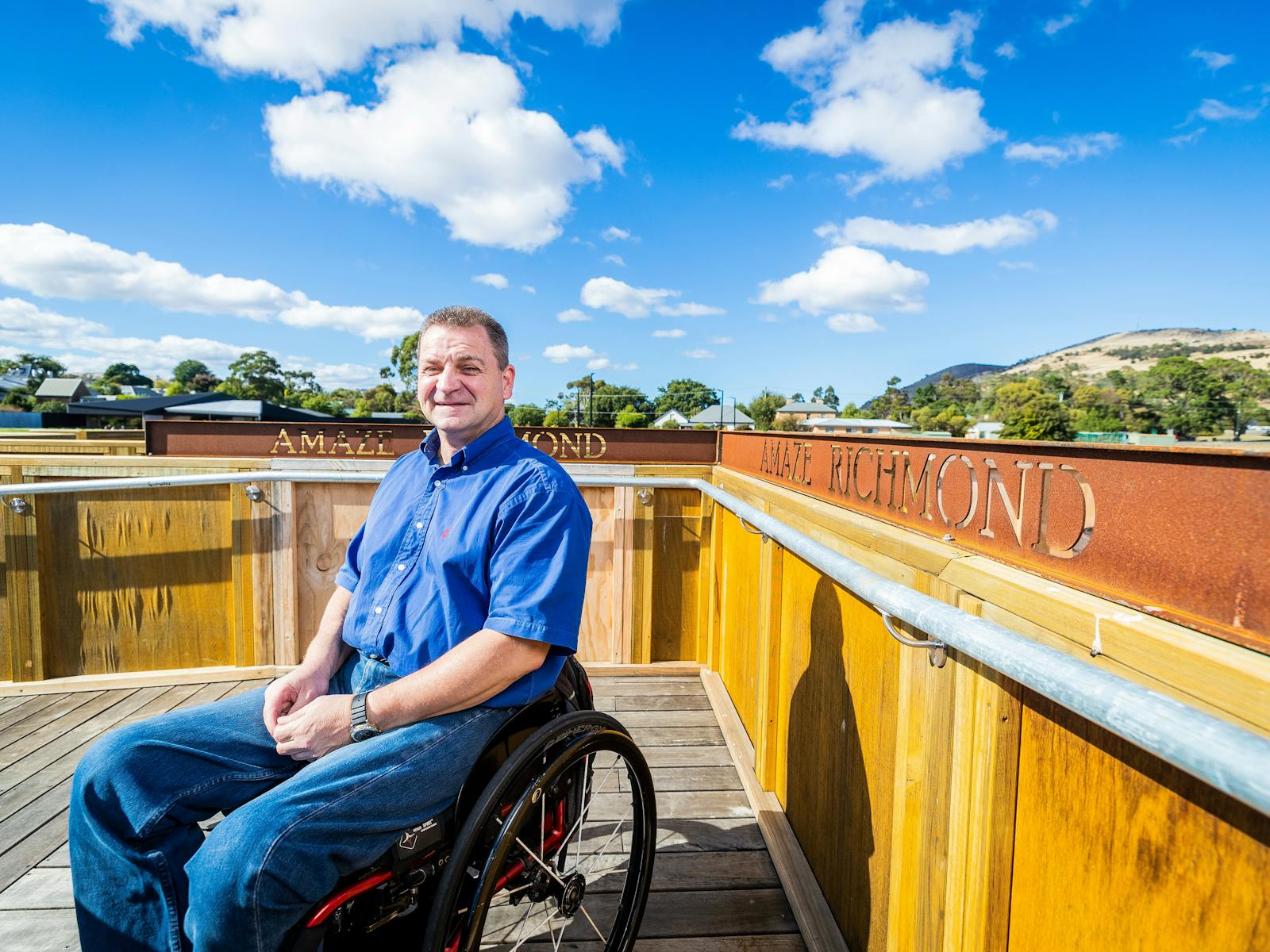 Smiling man wearing a blue shirt and jeans, sitting in a wheelchair in front of a metal sign.